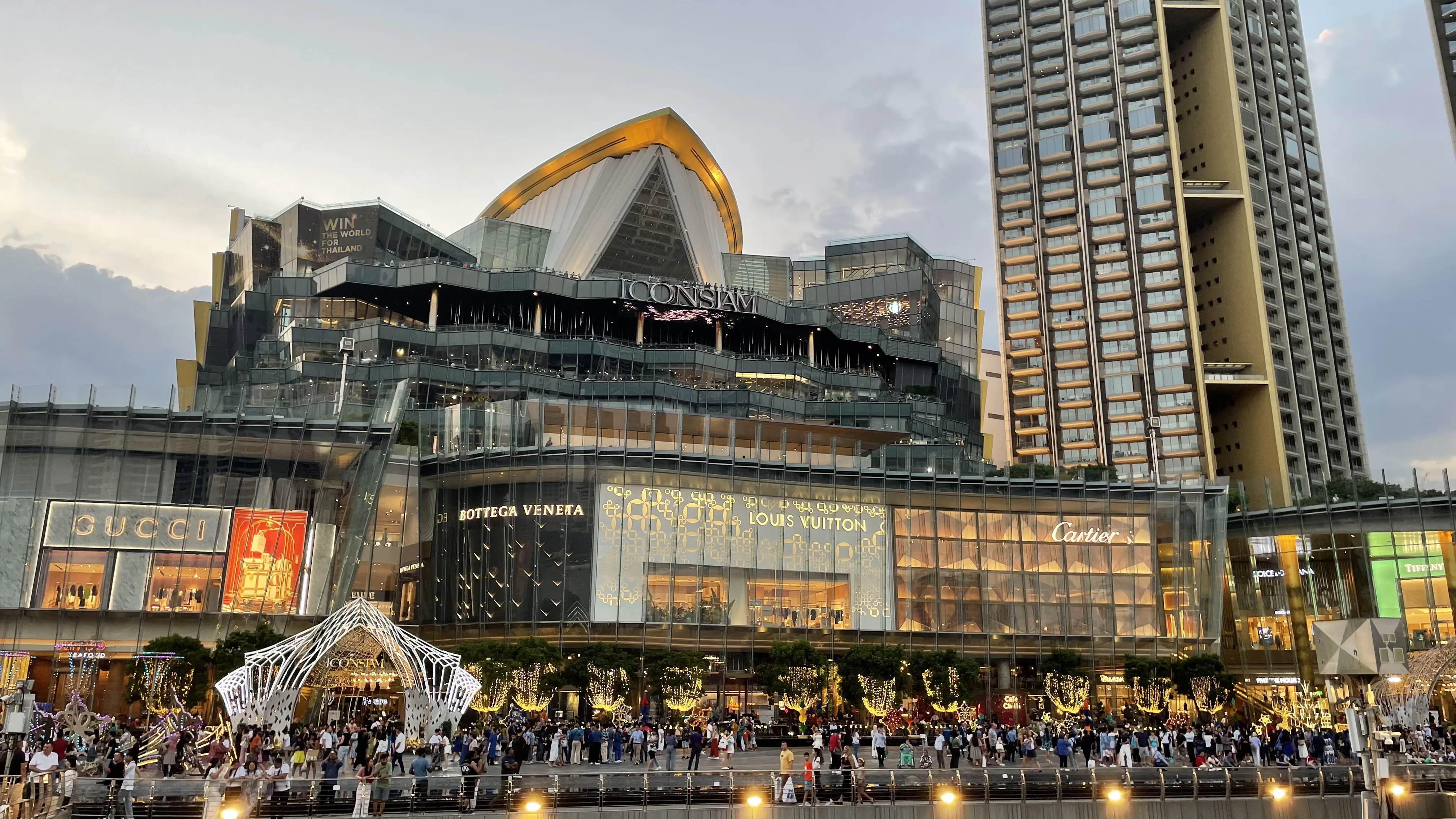 Vista externa do luxuoso complexo comercial ICONSIAM à noite, com lojas de grife iluminadas e grande movimento de pessoas na orla em Banguecoque.