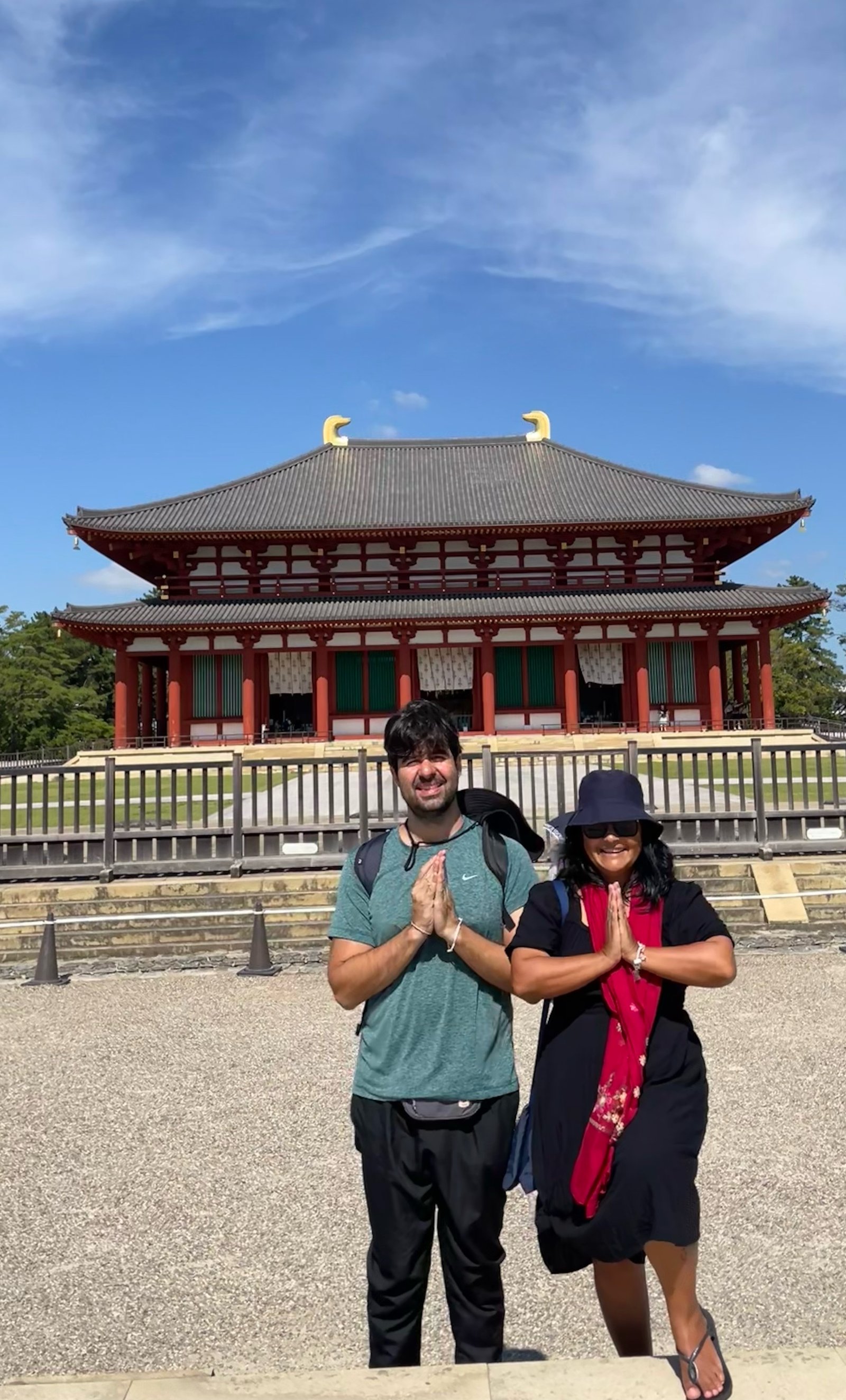Homem e mulher sorriem em posição de prece em frente ao templo budista Chu-kondo, de arquitetura vermelha e telhado duplo.
