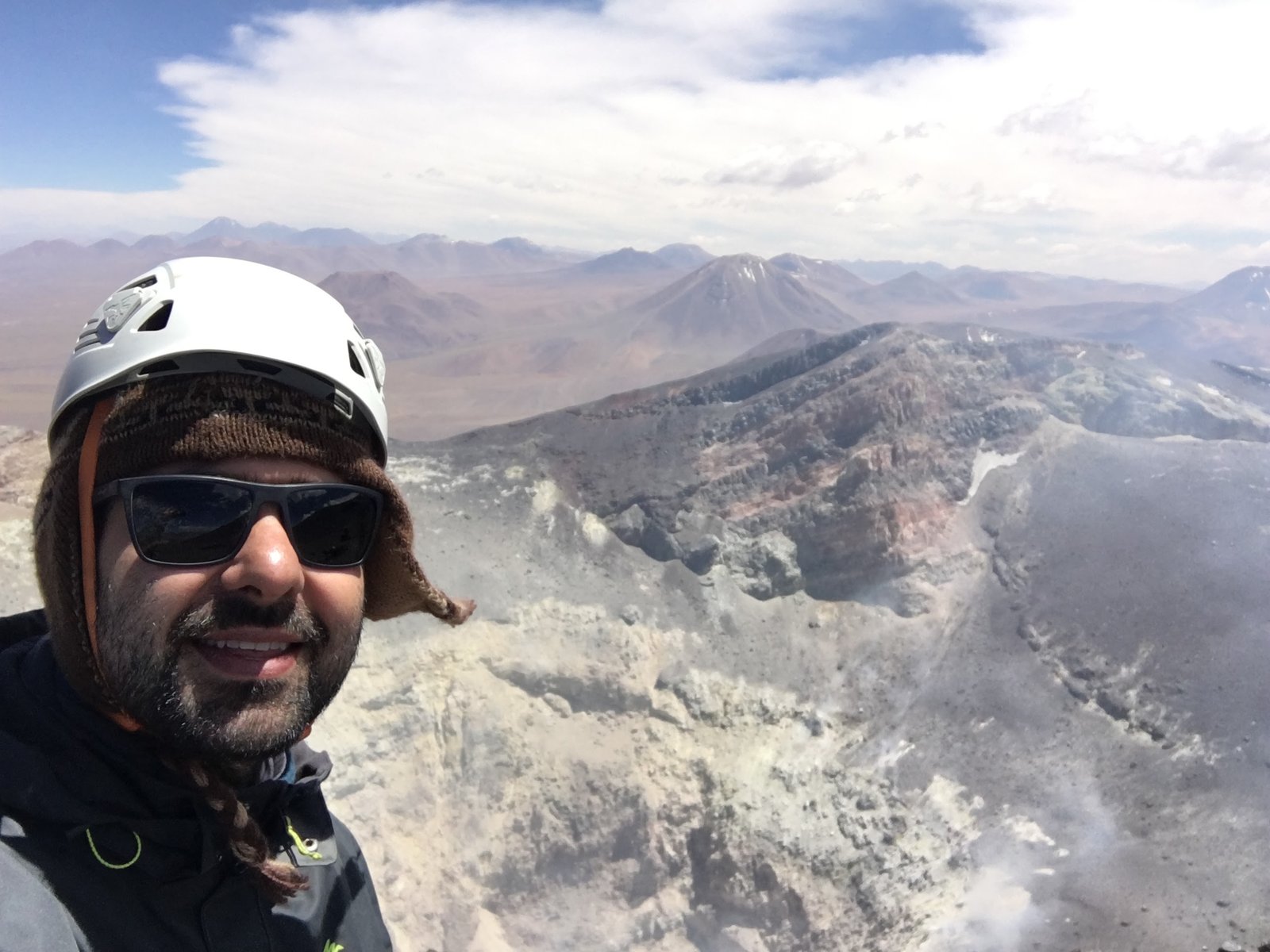 Homem de capacete e óculos de sol tira selfie com a cratera do Vulcão Lascar, no Atacama, ao fundo.