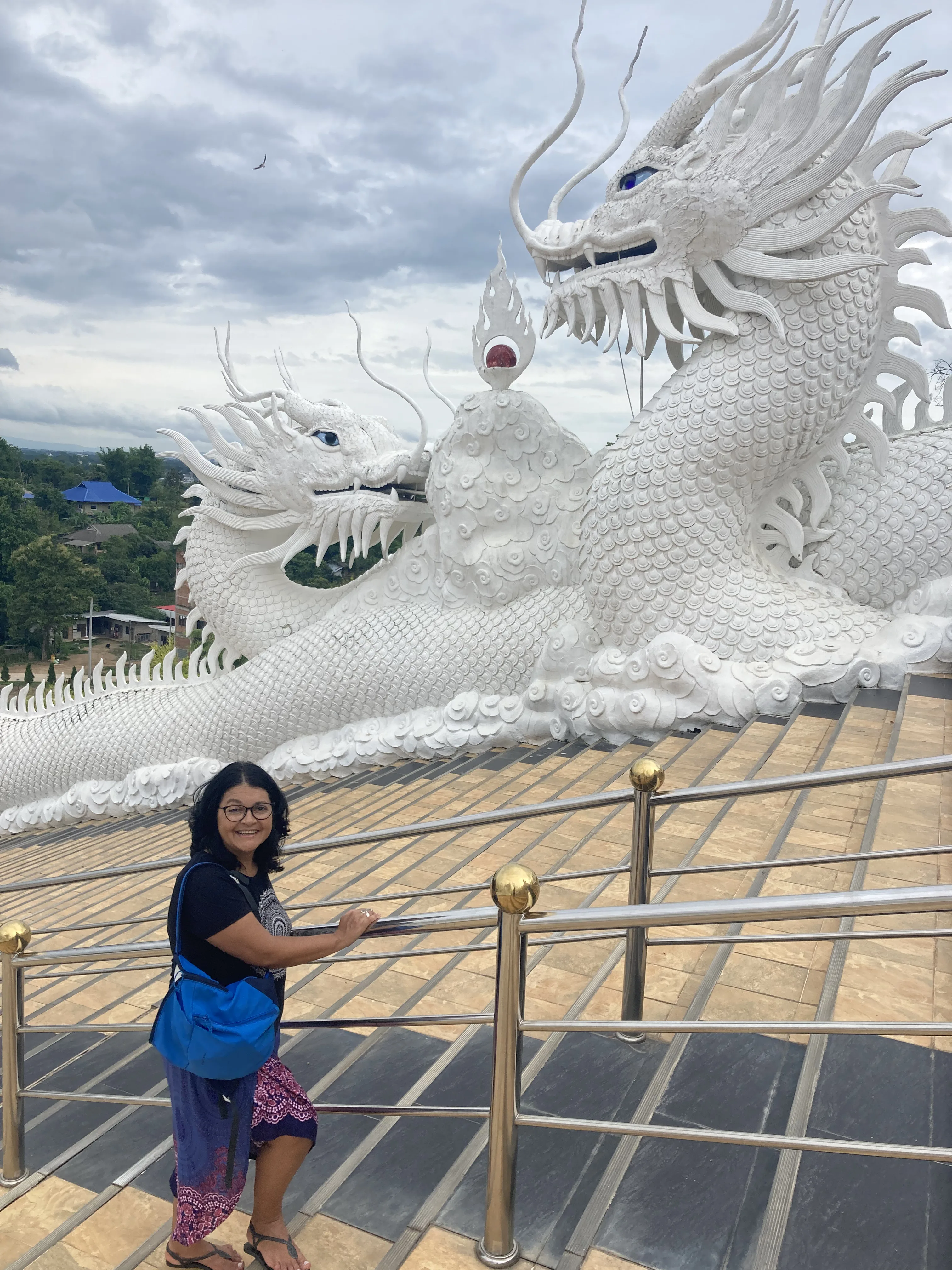 Uma mulher sorridente com uma bolsa azul em pé nos degraus ornamentados com grandes esculturas de dragões brancos na escadaria do templo em Chiang Rai