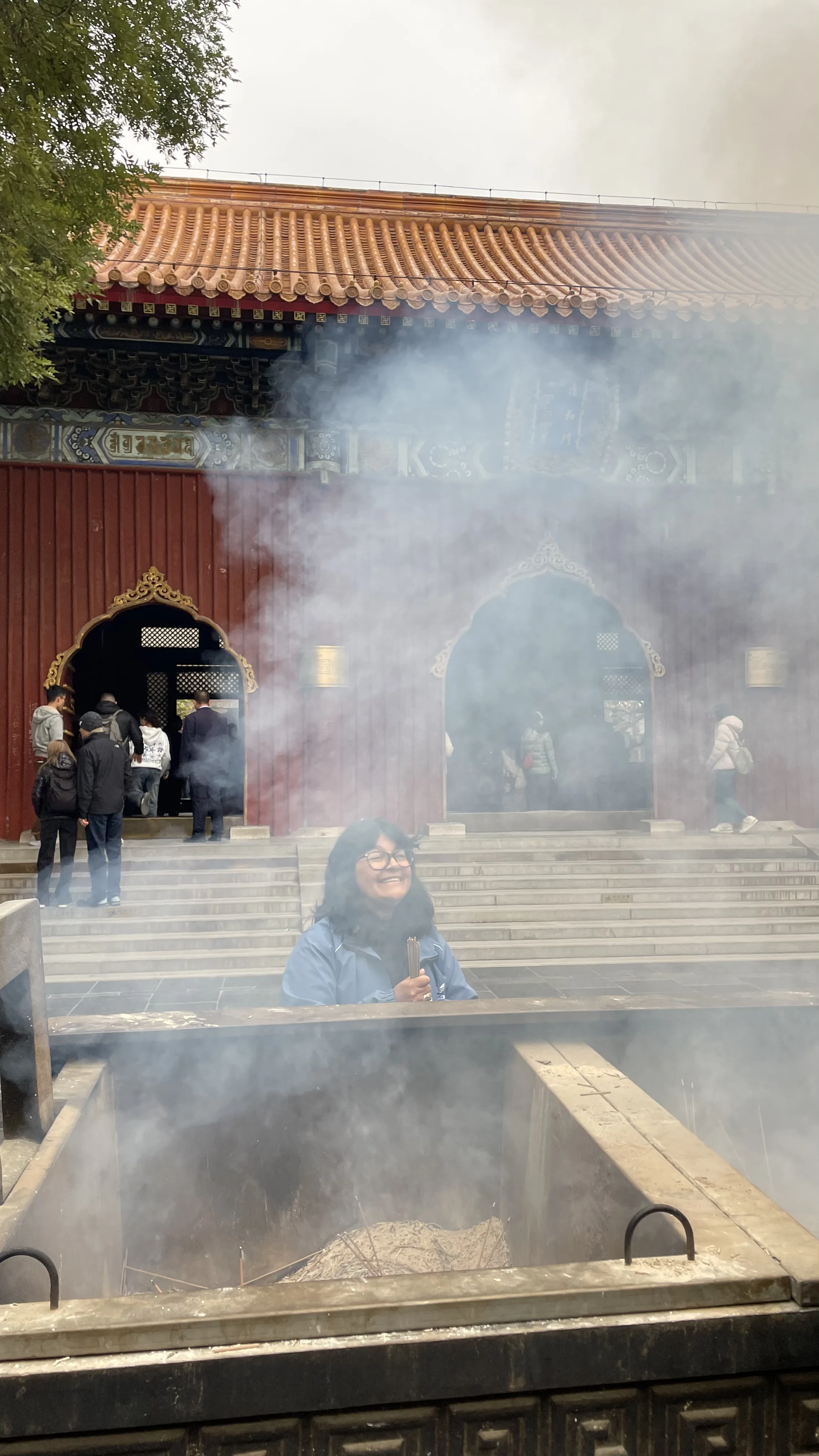 Mulher sorri com incensos nas mãos atrás de grande incensário metálico tomado por fumaça em pátio de templo em Pequim.