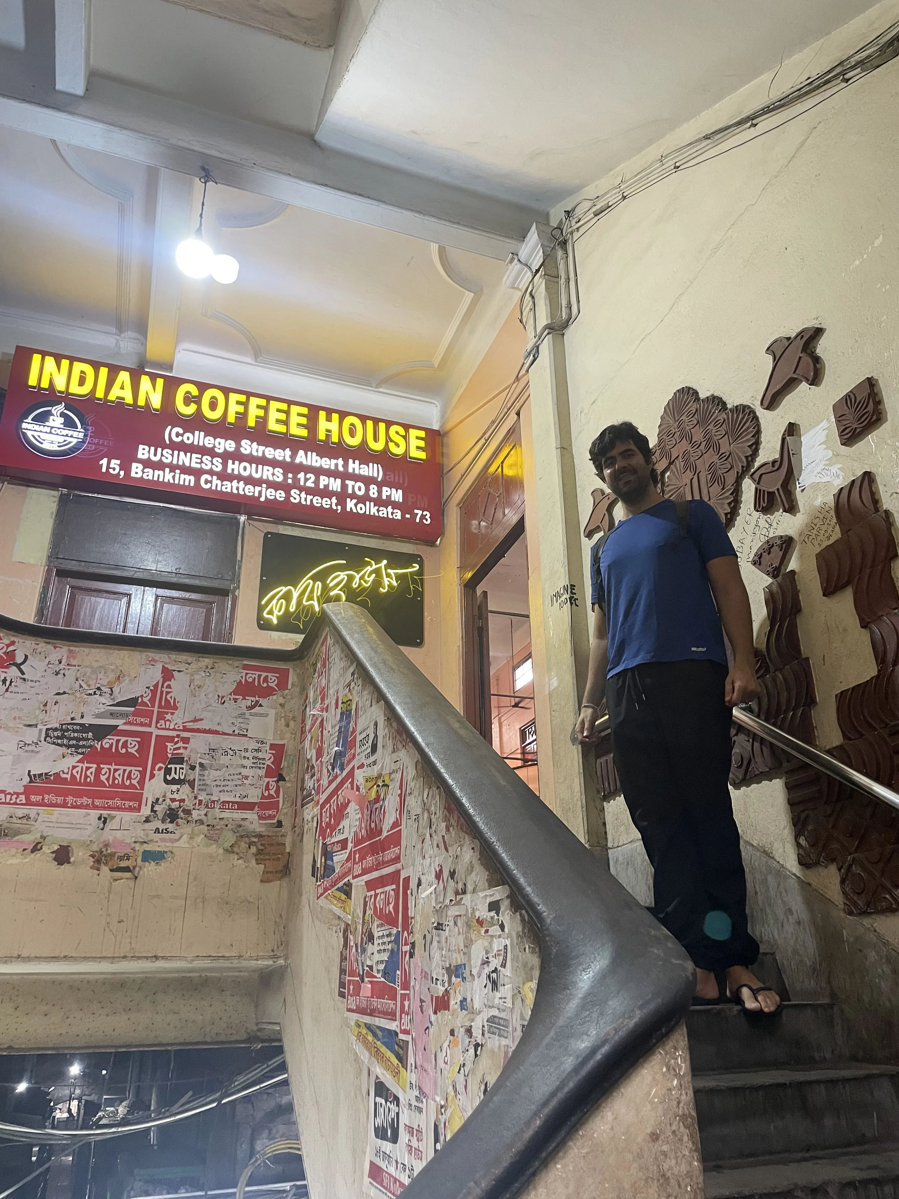 homem em pé sorrindo na escadaria interna do histórico Indian Coffee House em Calcutá, com placa vermelha iluminada na parede e cartazes colados nos arredores.