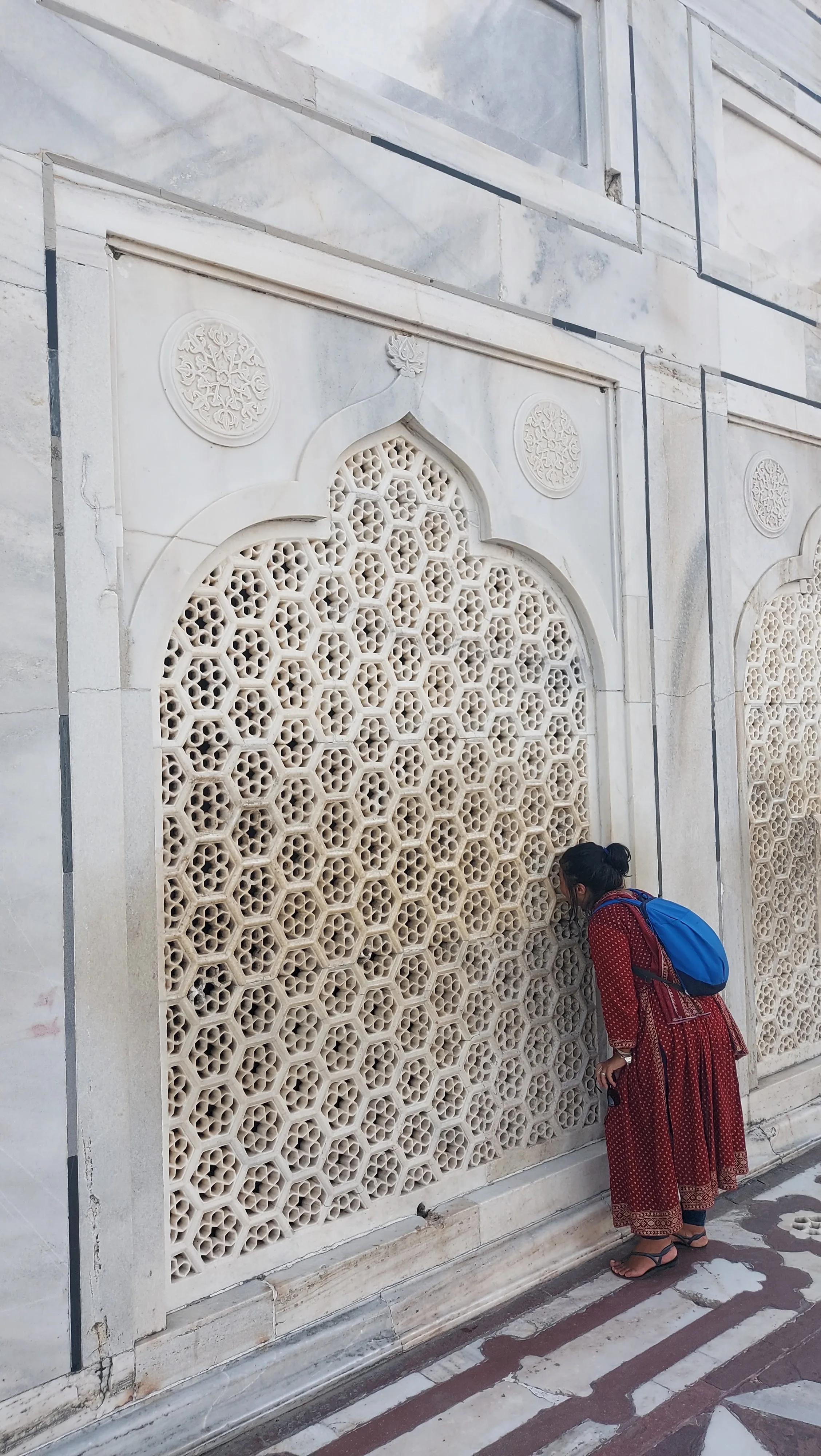 Visitante com vestido vermelho e mochila azul observando de perto uma janela celosia de mármore branco entalhada com padrões geométricos vazados em formato de favo de mel na parede externa do Taj Mahal.