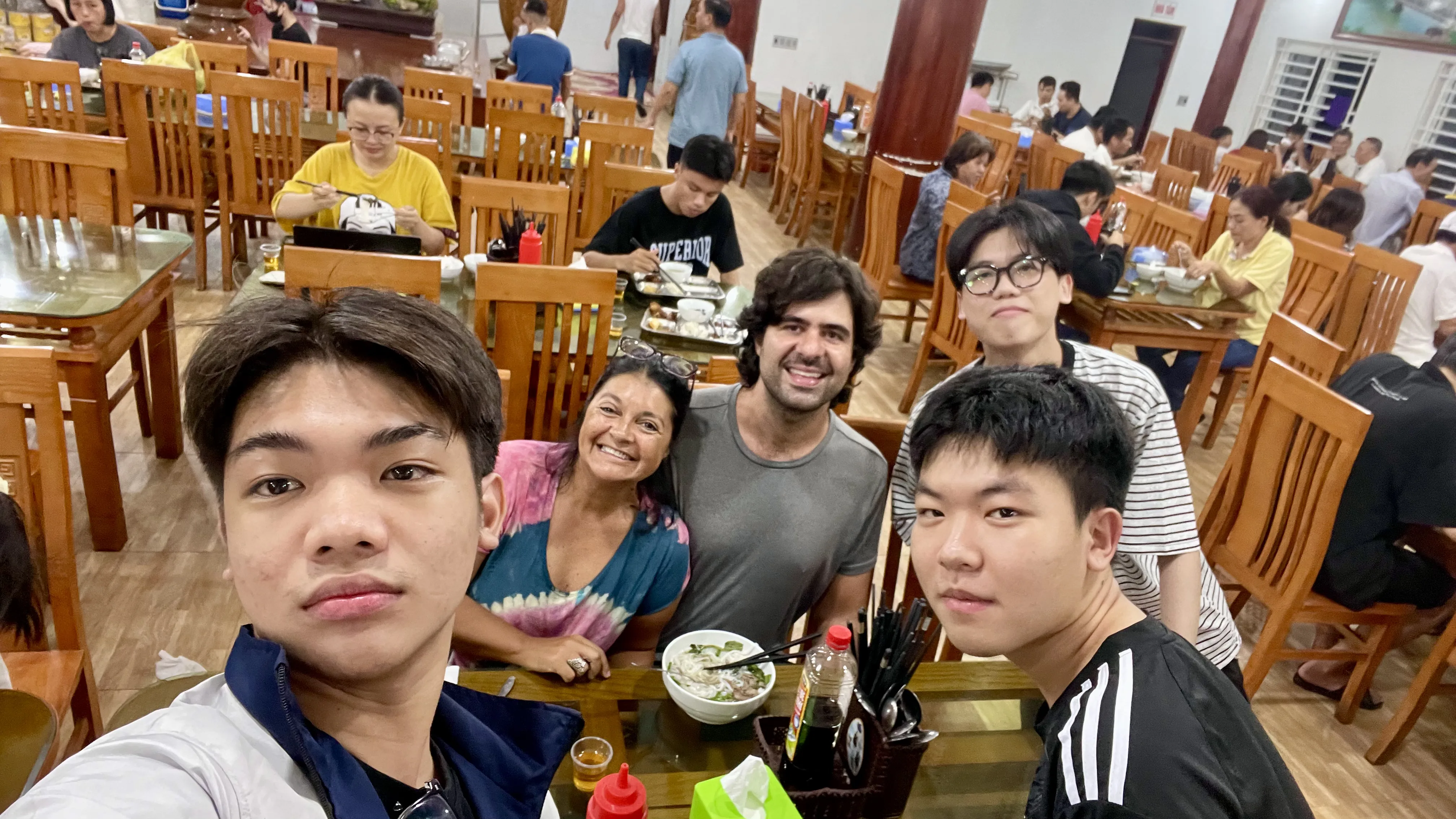 Grupo de passageiros a sorrir para uma selfie durante uma paragem para refeição num restaurante tradicional ao longo da viagem de autocarro no Vietname