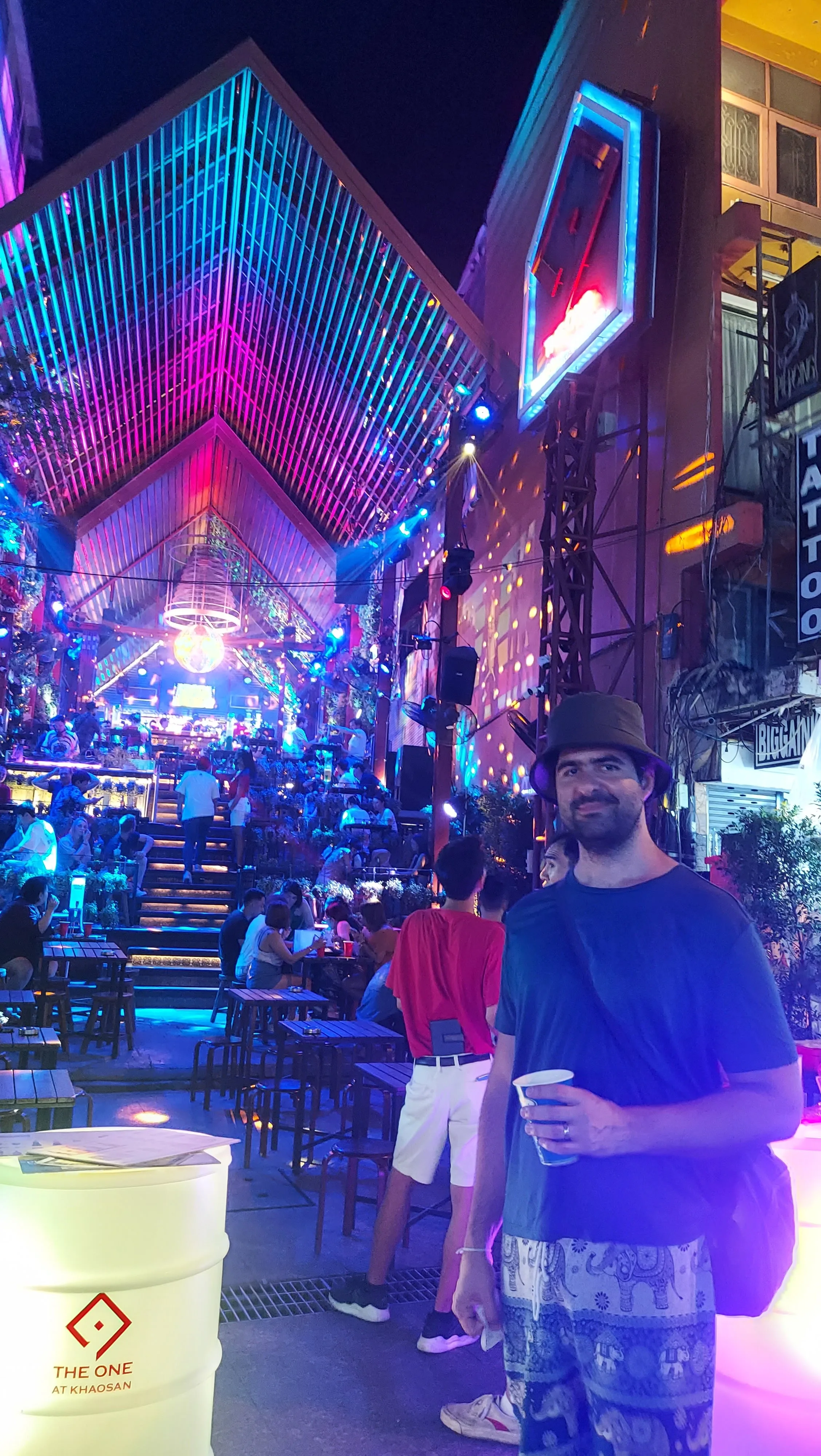 Homem sorridente segurando um copo à noite na vibrante Khaosan Road, iluminada por luzes neon e com o ambiente animado do The One at Khaosan ao fundo, em Banguecoque.