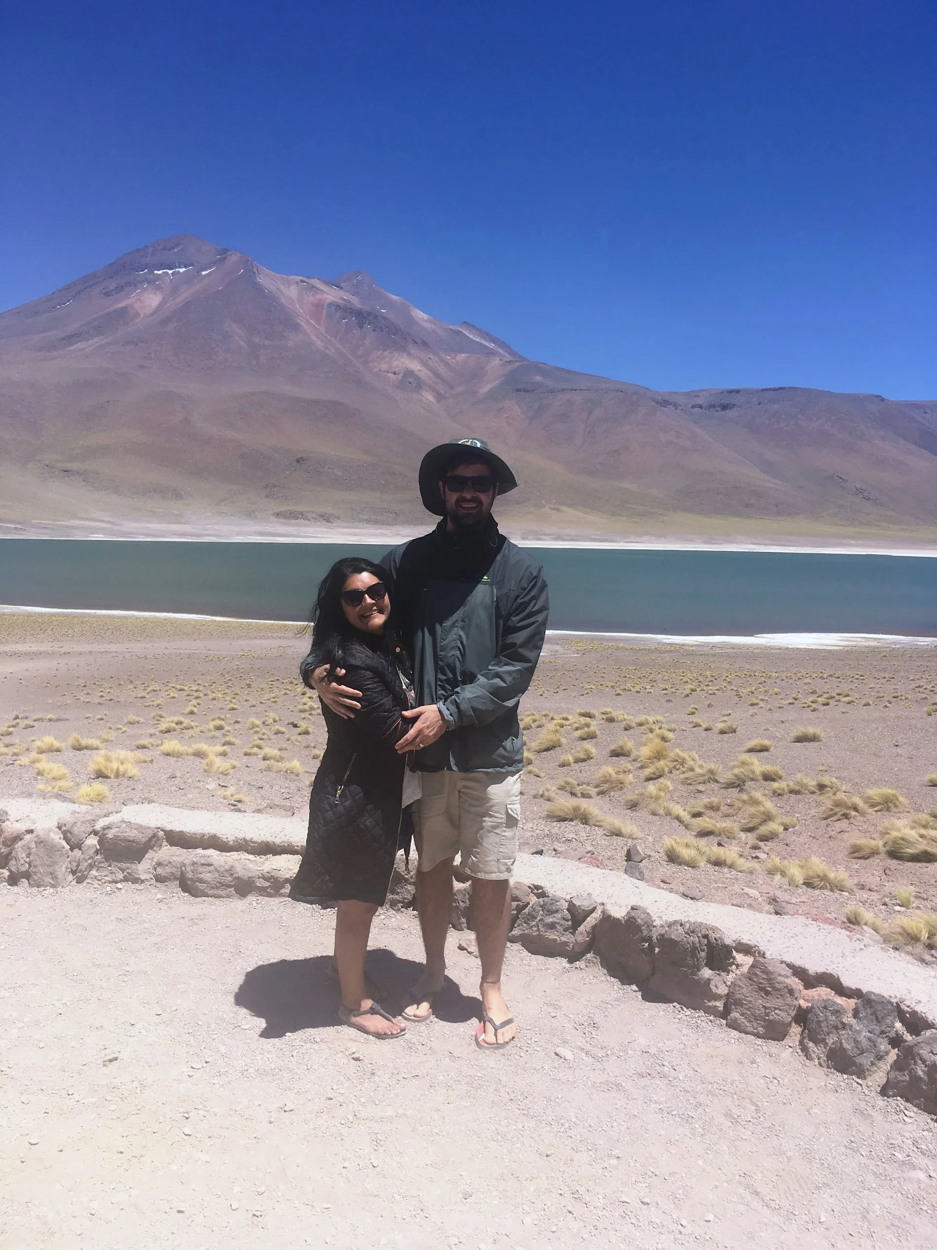 Casal abraçado sorri diante das águas azuis e vegetação rasteira com montanha ao fundo na bela Laguna Altiplânica no Chile.
