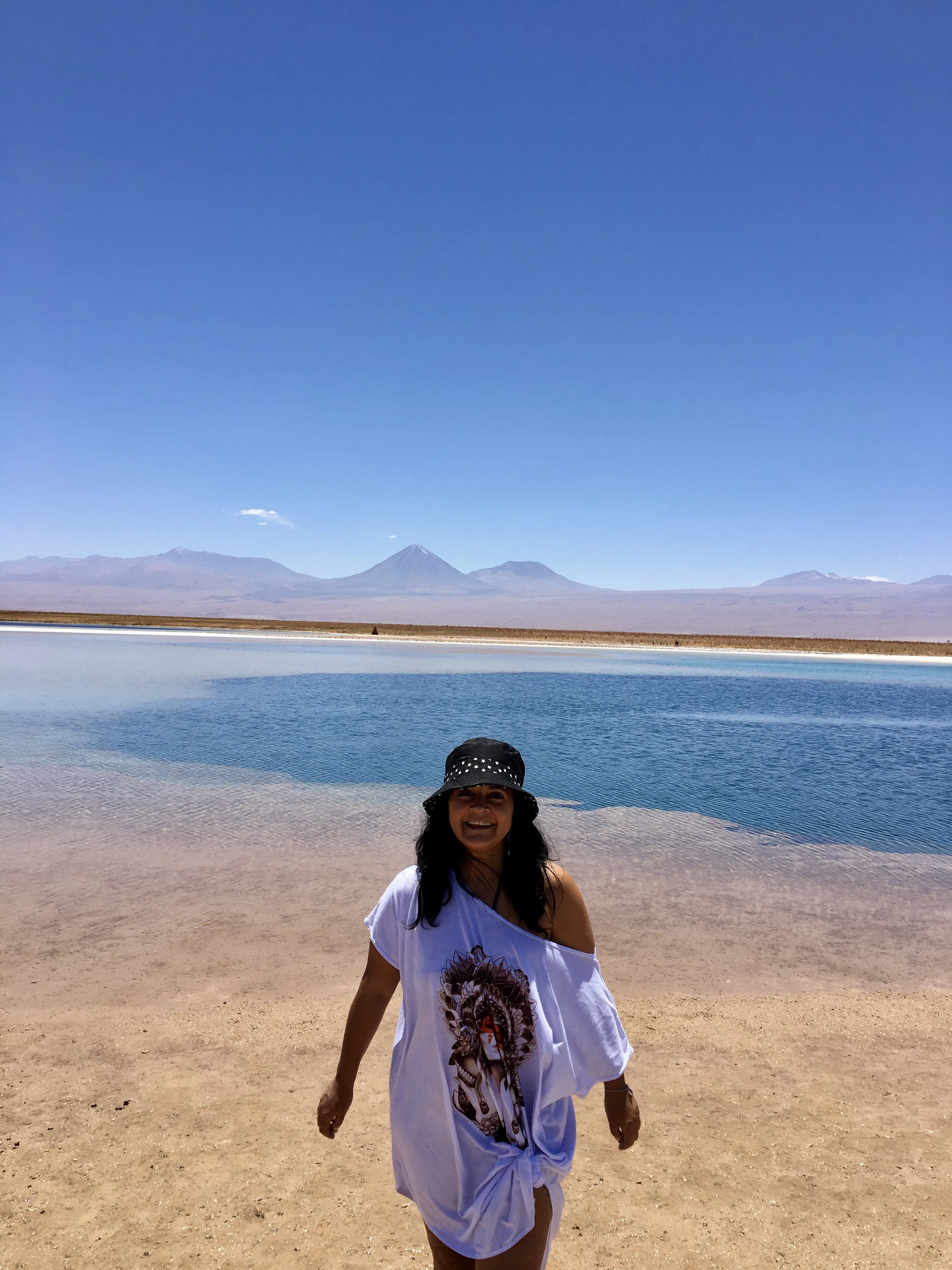 Mulher sorri na margem de areia clara diante das águas azuis e montanhas ao fundo na paisagem da Laguna Piedra no Atacama.