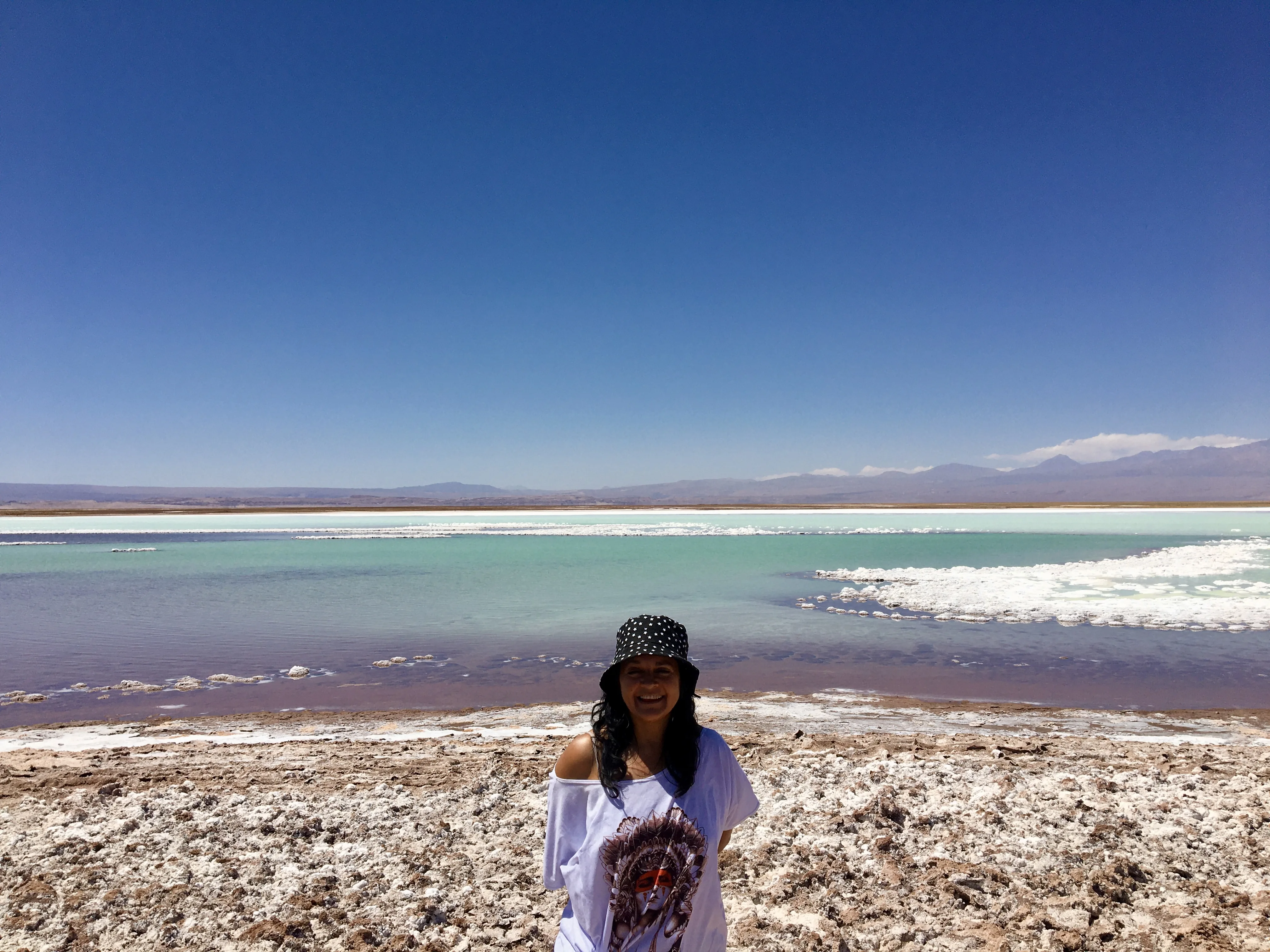 Mulher sorri em pé diante das águas turquesas com bordas de sal branco e montanhas ao fundo na Laguna Tebenquiche no Chile.