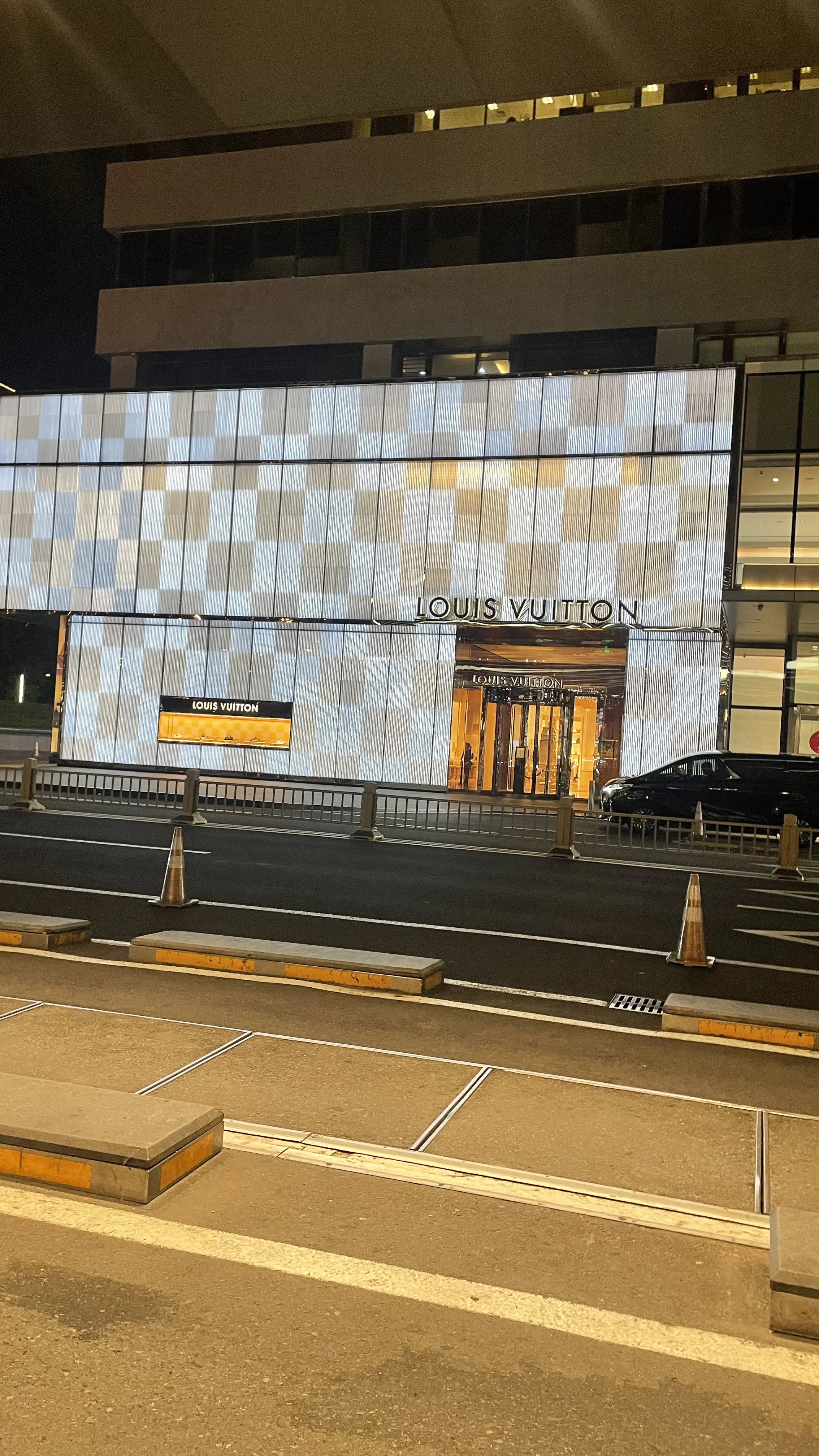 Fachada iluminada da loja Louis Vuitton à noite com vitrine quadriculada e rua asfaltada com cones de trânsito em Pequim.