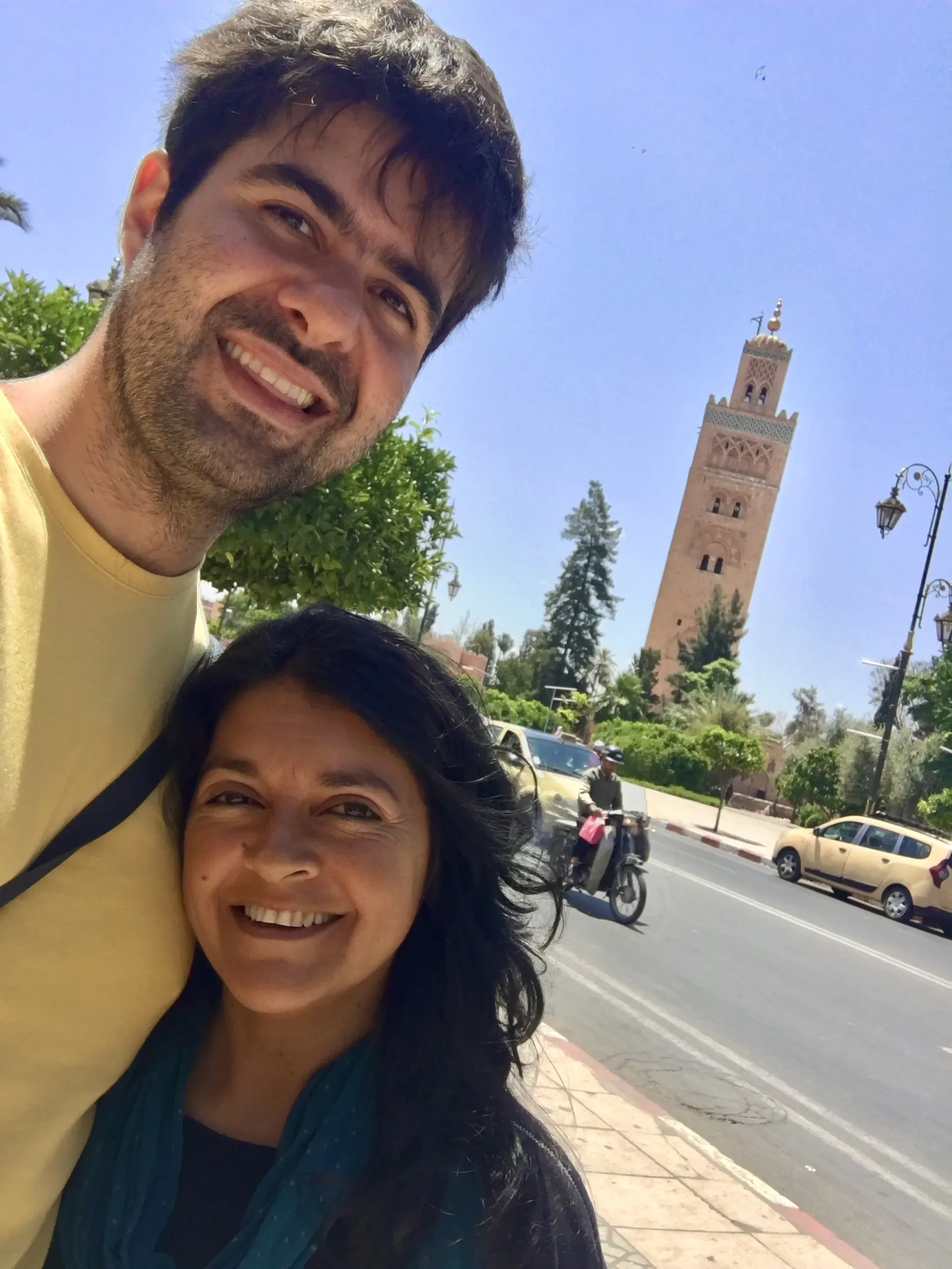 Casal sorri em rua de Marraquexe com a torre da Mesquita Koutoubia e carros ao fundo sob o céu azul.