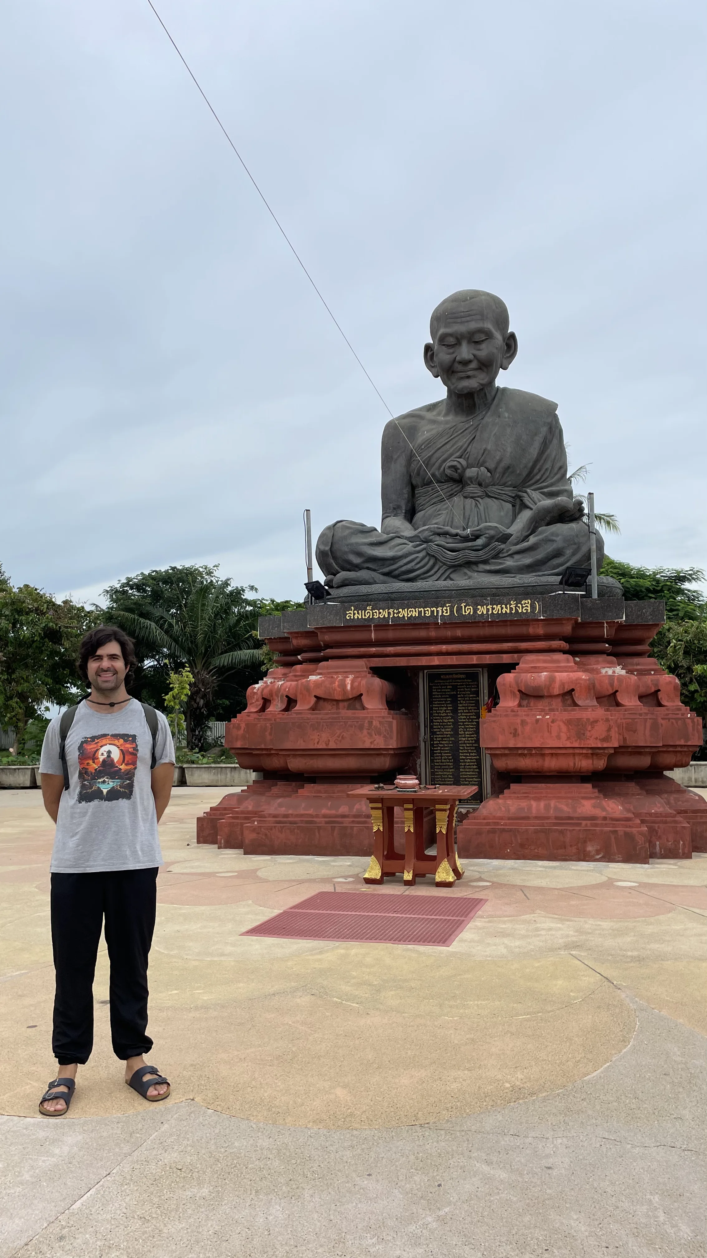 Turista posando ao lado da enorme estátua de bronze do venerado monge Somdej Toh, sentado em meditação sobre um grande pedestal vermelho ornamentado, no pátio ao ar livre de Wat Khun Inthapramun.