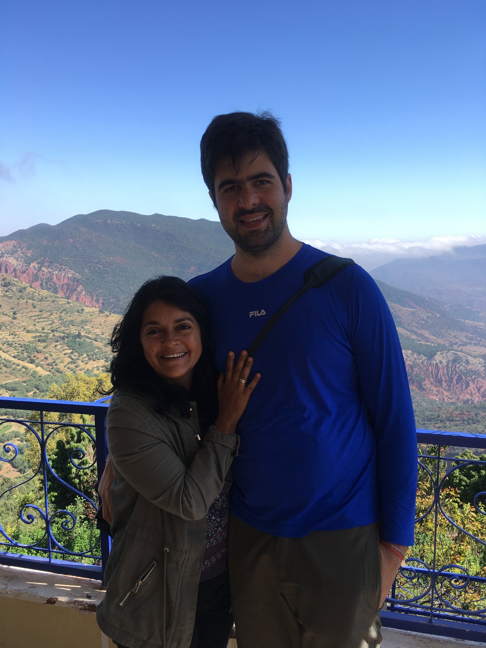 Casal sorri junto a parapeito azul com vista panorâmica para as Montanhas do Atlas em Marrocos.