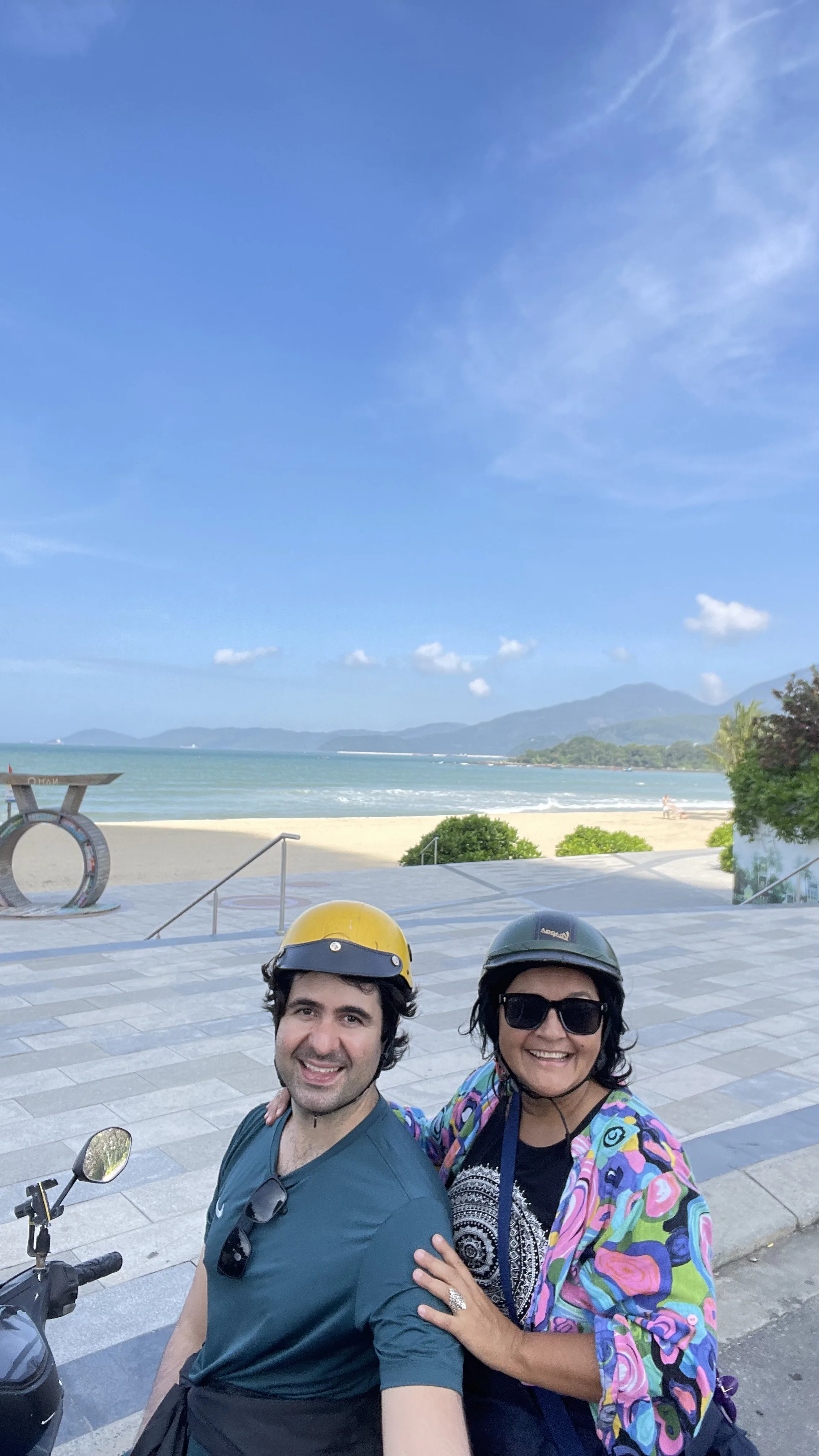 Selfie de casal sorridente de capacete e óculos de sol à beira-mar em Đà Nẵng, com a praia e montanhas ao fundo.