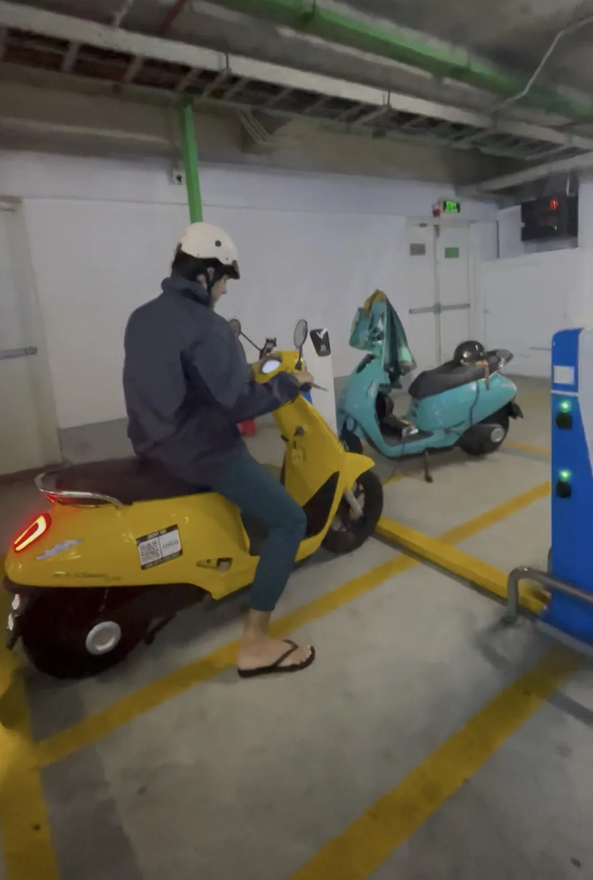 Pessoa a conduzir uma scooter elétrica amarela numa garagem subterrânea equipada com pontos de carregamento.