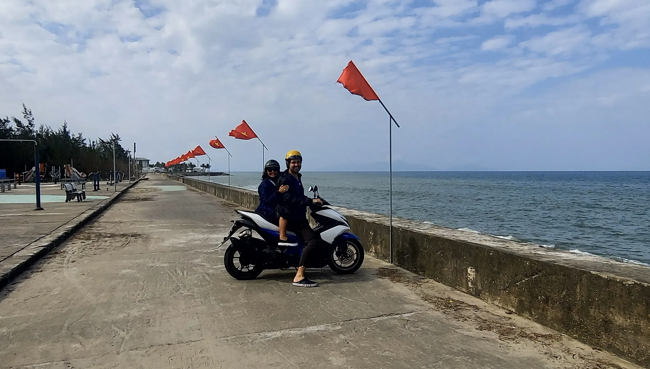 Casal de capacete numa scooter junto a um paredão costeiro enfeitado com pequenas bandeiras vermelhas em Hoi An.