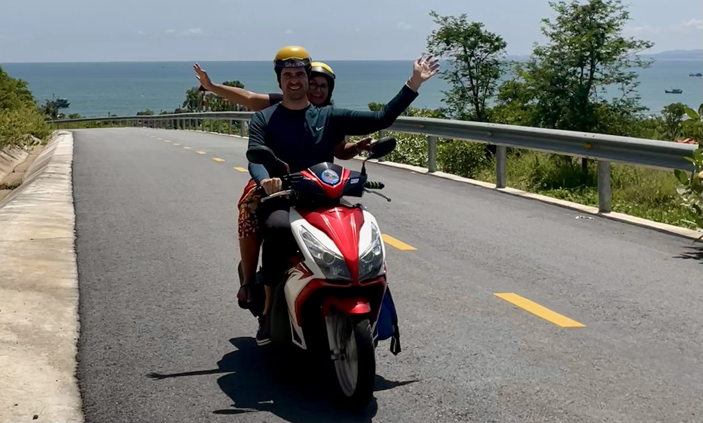 Casal sorridente a passear de moto com os braços abertos numa estrada costeira em Mũi Né, com o mar azul ao fundo.