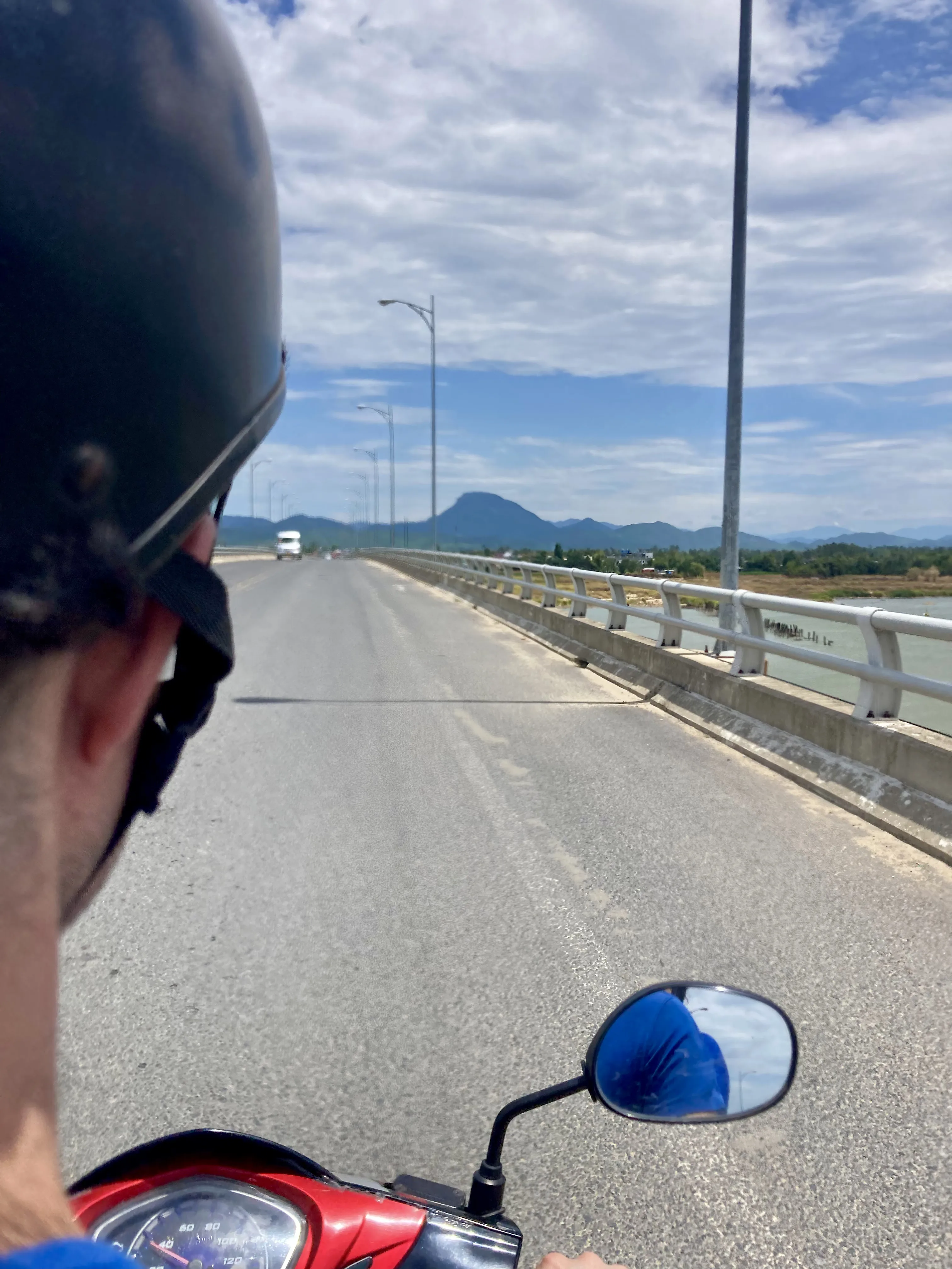 Vista em primeira pessoa do passageiro de uma motocicleta usando capacete, olhando para uma longa ponte sobre um rio em direção a montanhas distantes sob um céu azul com nuvens.