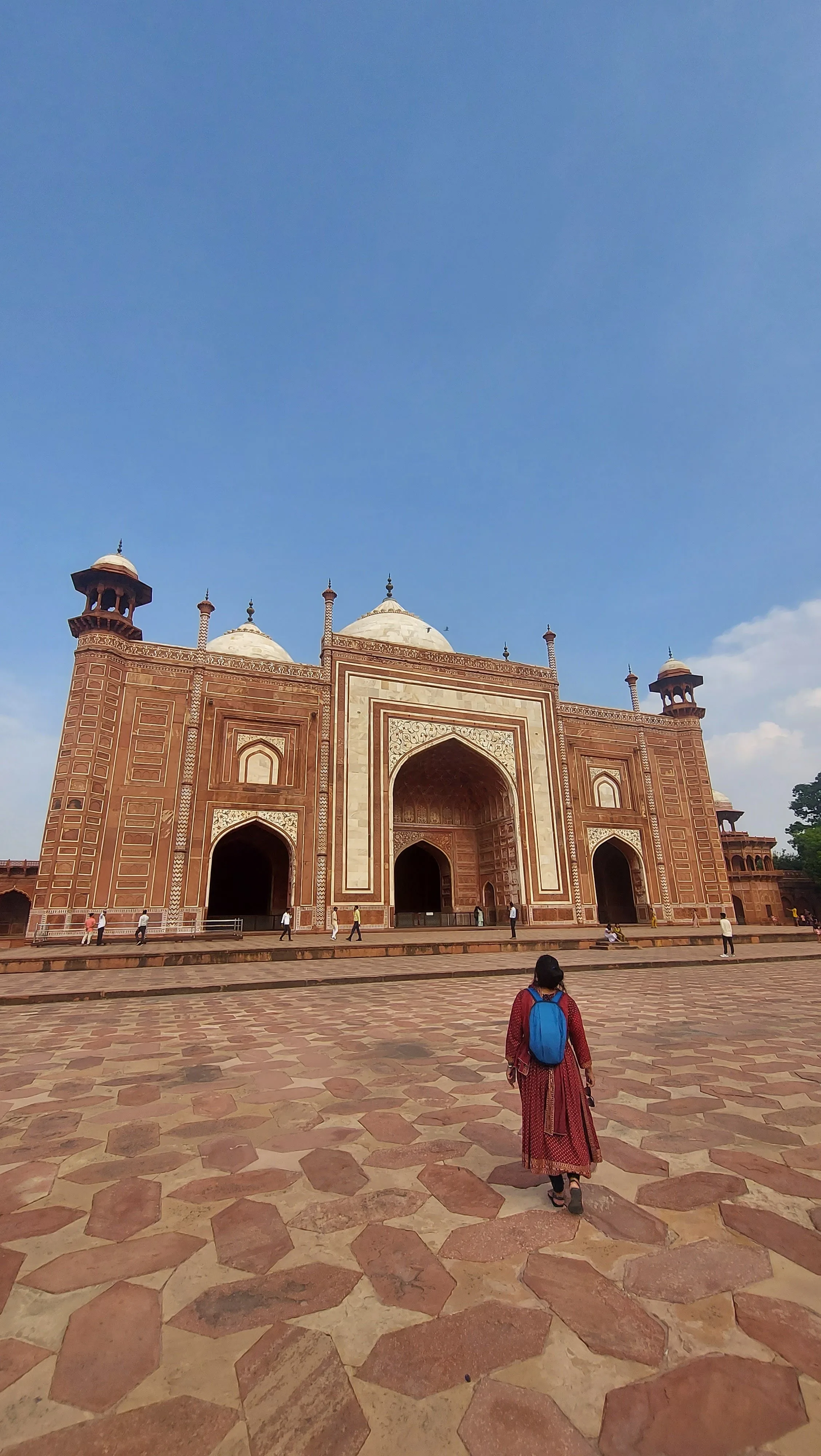 Visitante com vestido vermelho e mochila azul vista de costas caminhando pelo pátio de lajotas de arenito vermelho, em direção à imponente fachada de arenito vermelho e detalhes em mármore branco da mesquita lateral do complexo do Taj Mahal sob um céu azul.