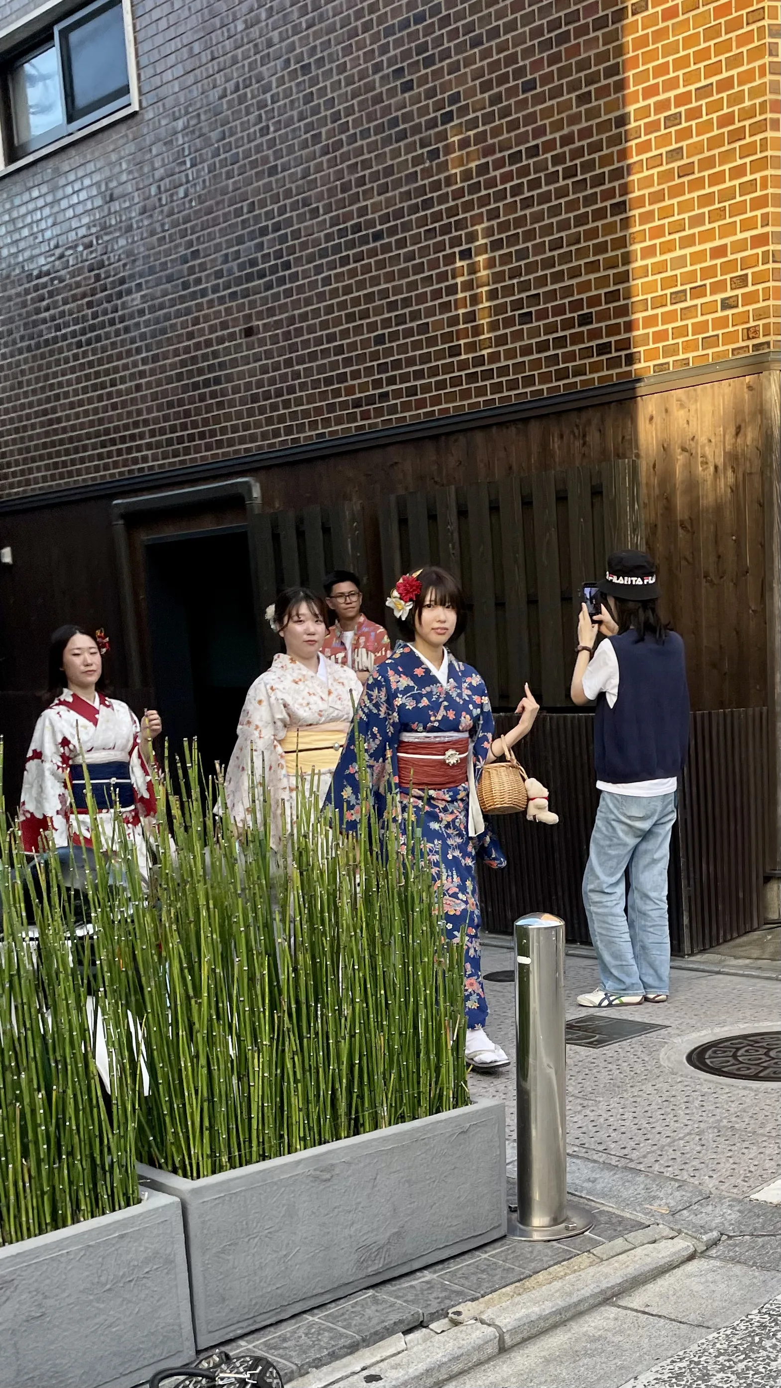 Três mulheres vestindo quimonos tradicionais coloridos posam em uma rua pavimentada do bairro de Gion, em Kyoto, enquanto outra pessoa as fotografa ao lado de vasos de plantas verdes.