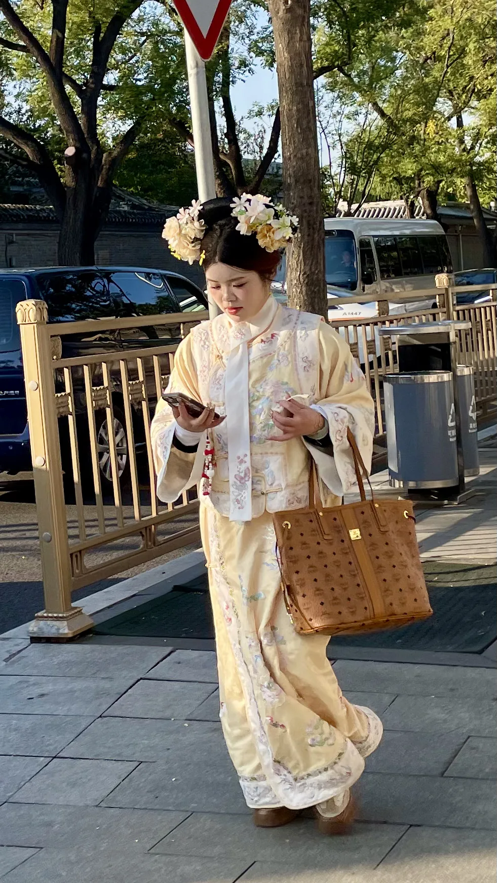 Mulher com traje tradicional amarelo e flores no cabelo caminha na calçada olhando o celular em Pequim.