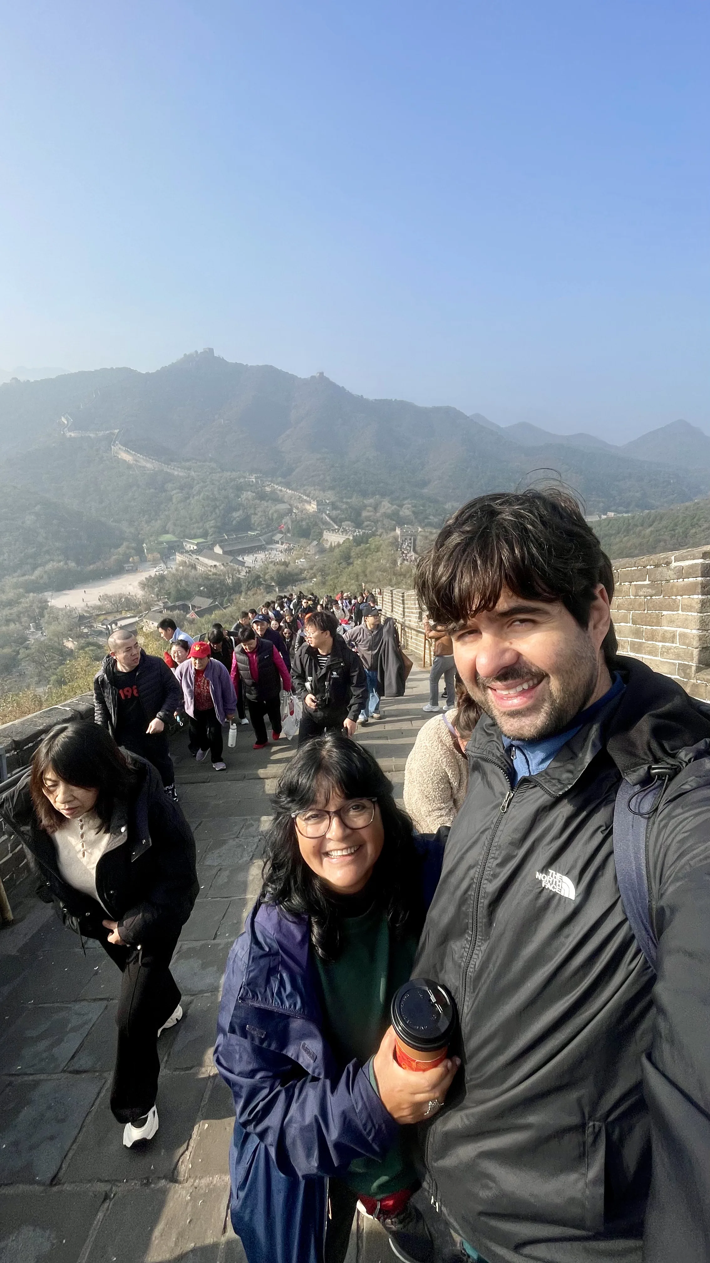 Uma selfie de um casal sorrindo em primeiro plano, segurando um copo de café, enquanto caminham em uma seção movimentada da Muralha da China, com uma longa fileira de outros turistas atrás deles e a icônica fortificação serpenteando por montanhas verdes sob um céu azul claro ao fundo.