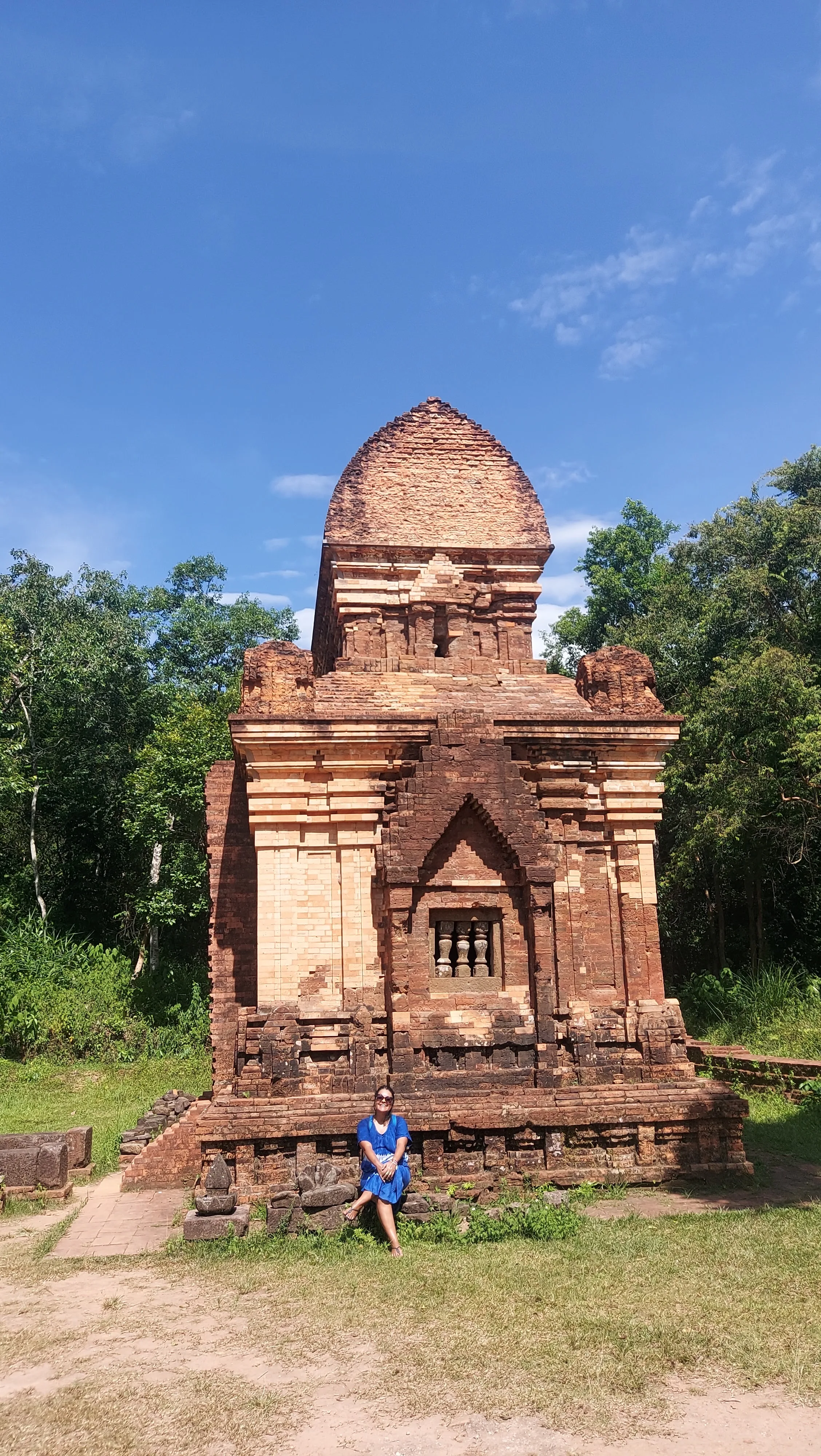 Mulher sentada perto das ruínas ancestrais e detalhadas de um templo de tijolos vermelhos no santuário histórico de My Son, no Vietname, rodeadas por vegetação sob um céu azul repleto de nuvens brancas.