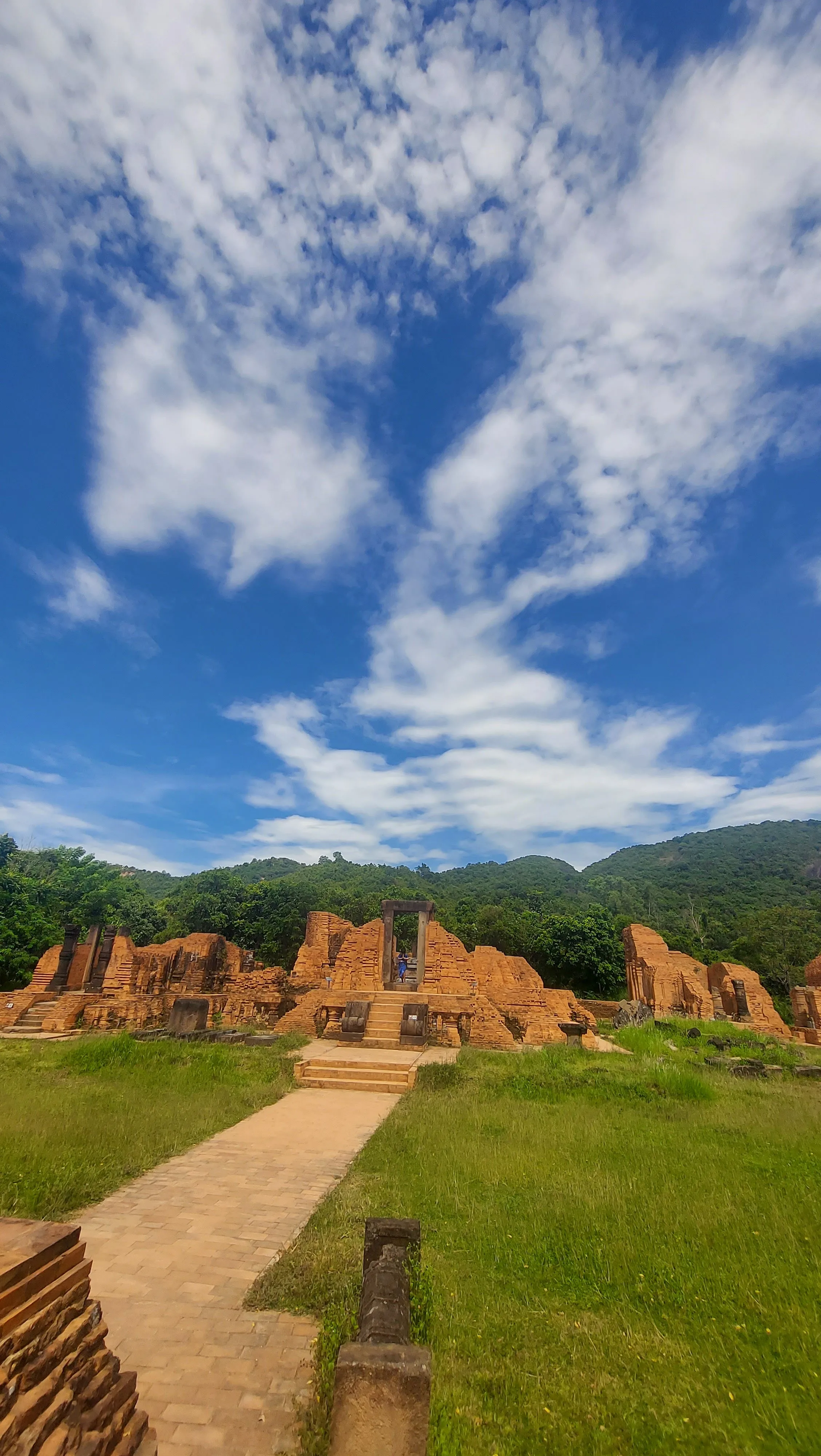 Ruínas ancestrais e detalhadas de um templo de tijolos vermelhos no santuário histórico de My Son, no Vietname, rodeadas por vegetação sob um céu azul repleto de nuvens brancas.