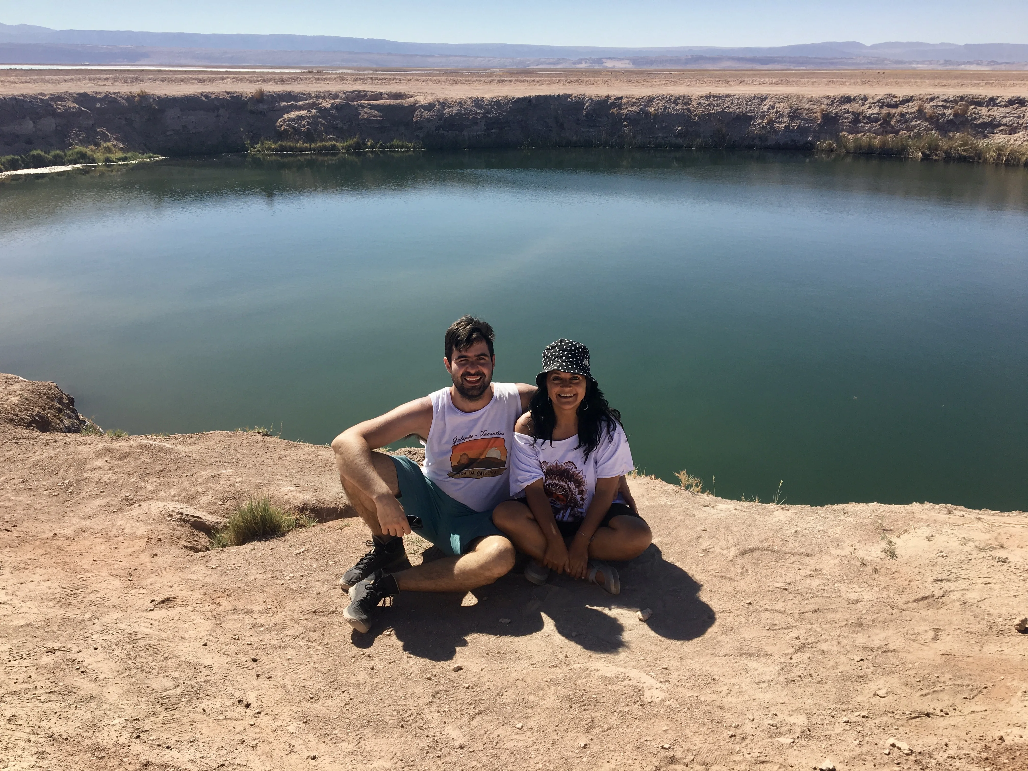 Casal sorri sentado na beira de terra diante das águas azuis da lagoa circular nos Ojos del Salar no Deserto do Atacama.