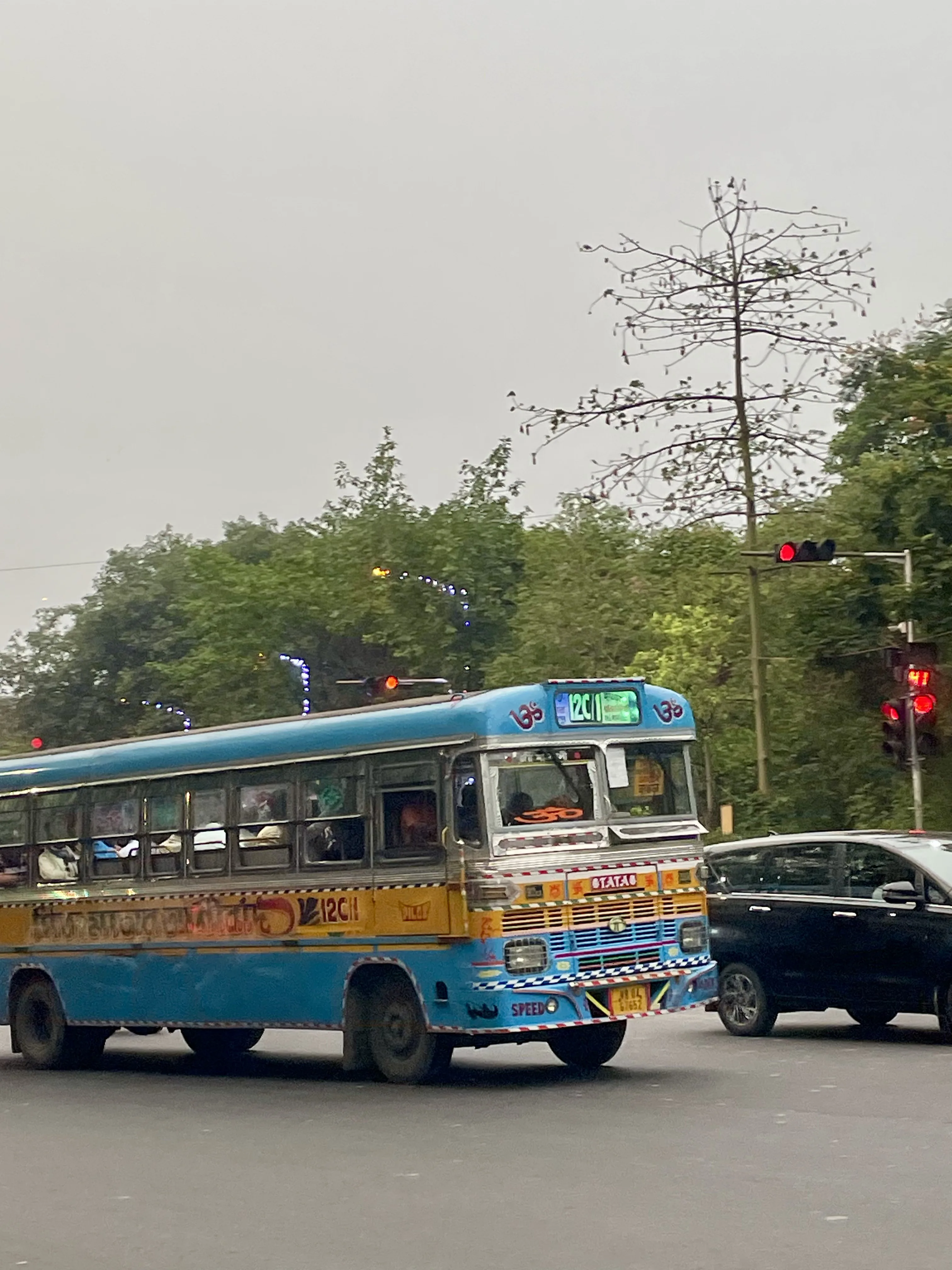 Ônibus de transporte público pintado em tons de azul e amarelo trafegando por uma rua movimentada em Calcutá, tendo ao fundo árvores frondosas e um semáforo com luz vermelha acesa.