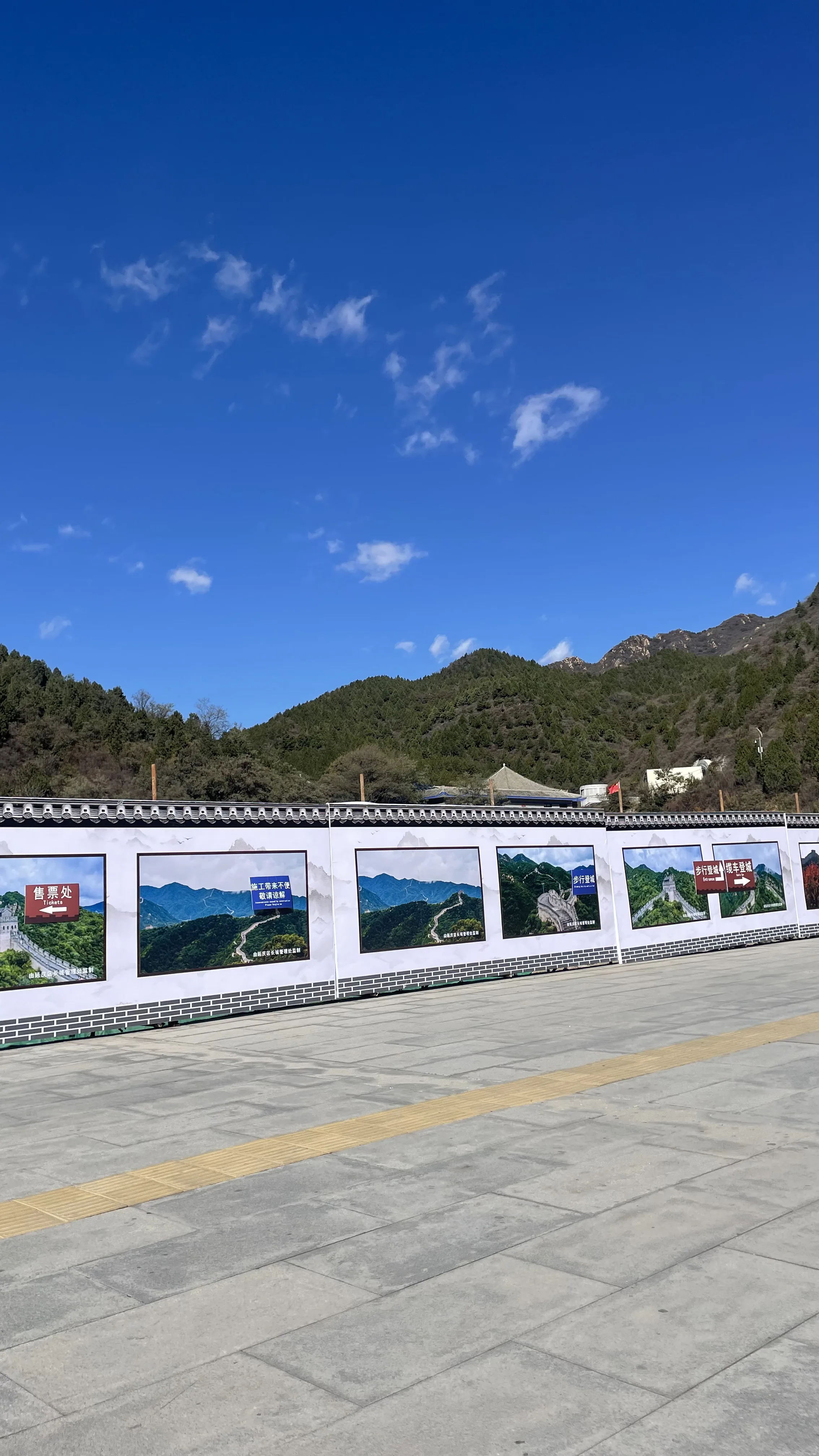 Painel decorativo com várias fotos da Muralha da China ao longo de montanhas cobertas por vegetação sob um céu azul brilhante com poucas nuvens, instalado em uma praça pavimentada.