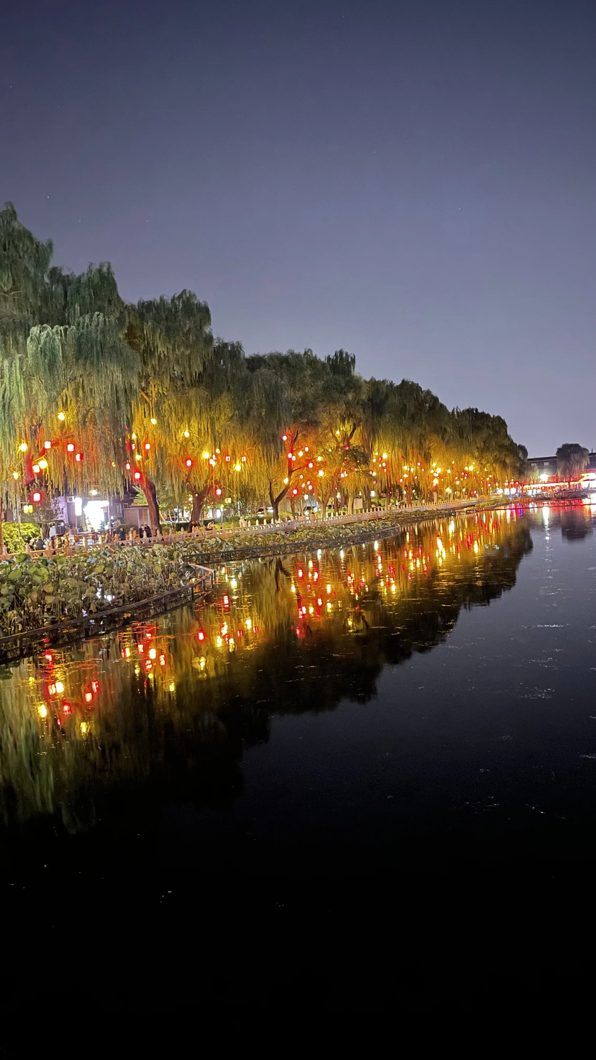 Ponte de pedra arqueada e árvores chorões iluminadas refletidas nas águas escuras de lago à noite em Pequim.