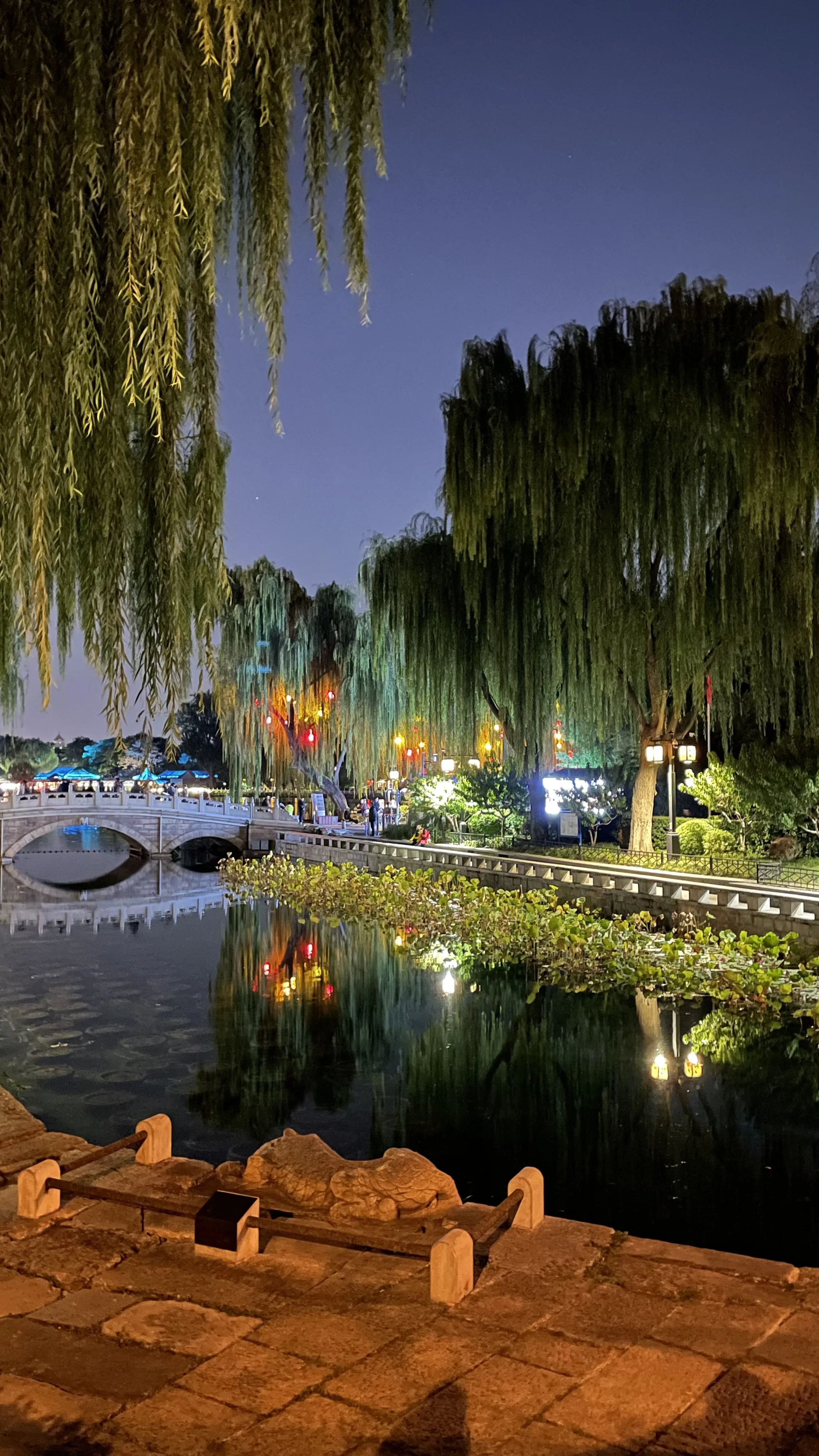 Ponte de pedra arqueada e árvores chorões iluminadas refletidas nas águas escuras de lago à noite em Pequim.