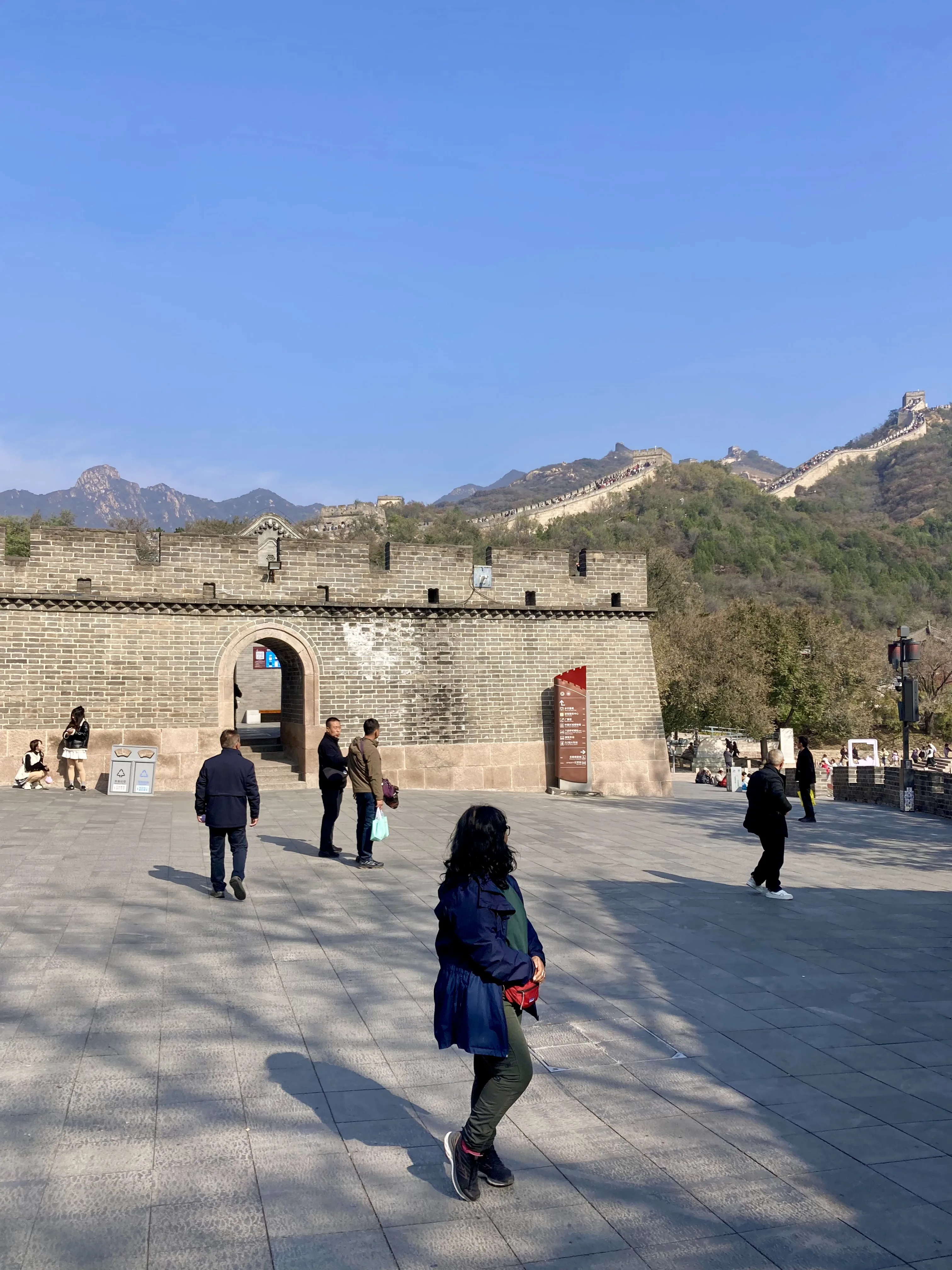 Turistas passeando no pátio de entrada pavimentado em frente ao portão em arco da muralha de tijolos de Badaling na Muralha da China, com a grande fortificação serpenteando pelas montanhas verdes ao longe sob um céu azul claro.