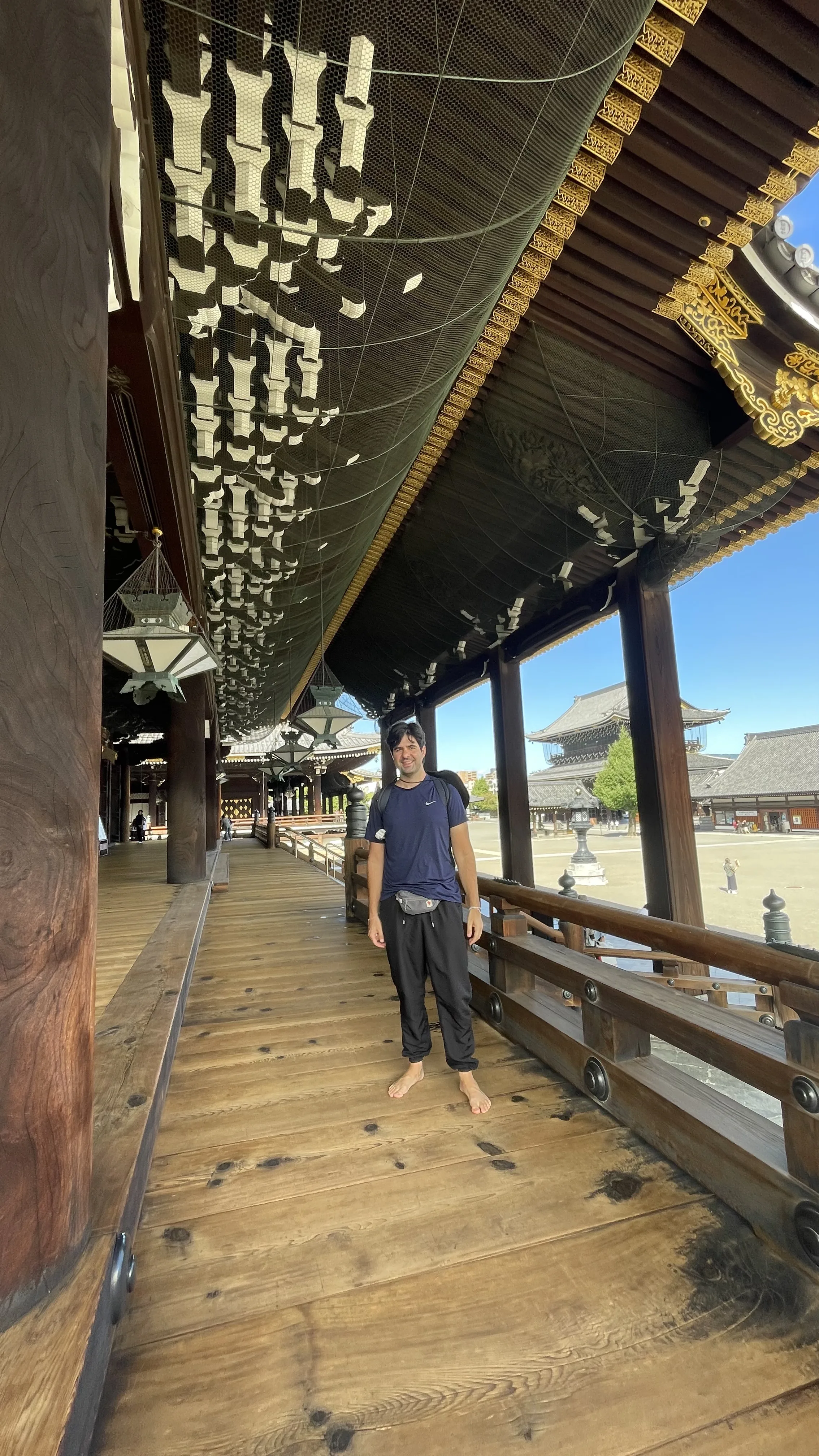 Um homem em pé, descalço, no Templo Hongan-ji em uma longa passarela de madeira polida com um teto ornamentado, sustentado por vigas de madeira escura. A estrutura tem uma grade de madeira baixa e vista para o pátio externo do templo, com outro edifício do templo ao longe.