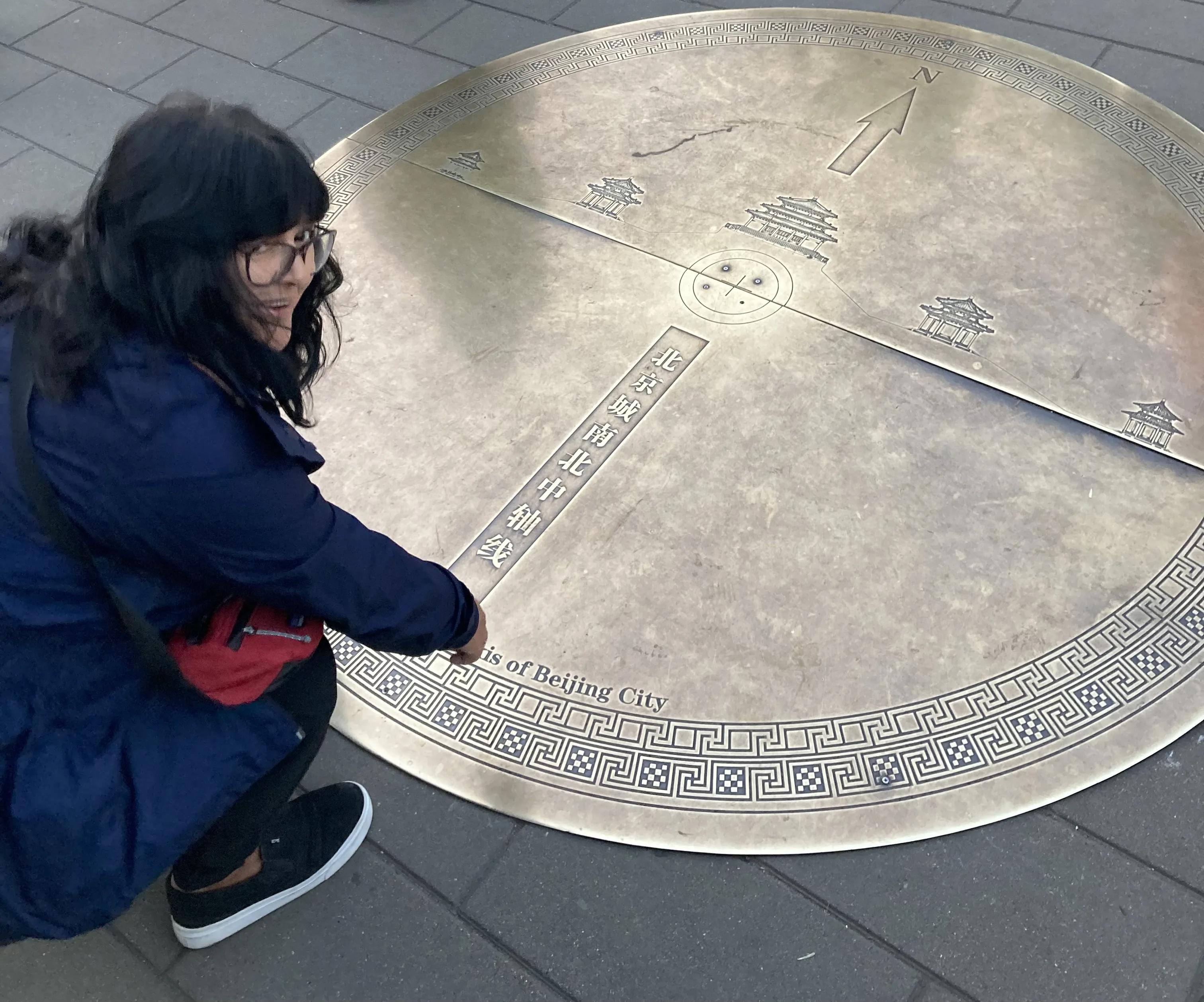 Mulher agachada aponta para placa metálica no chão com mapa e inscrições sobre o eixo central de Pequim.