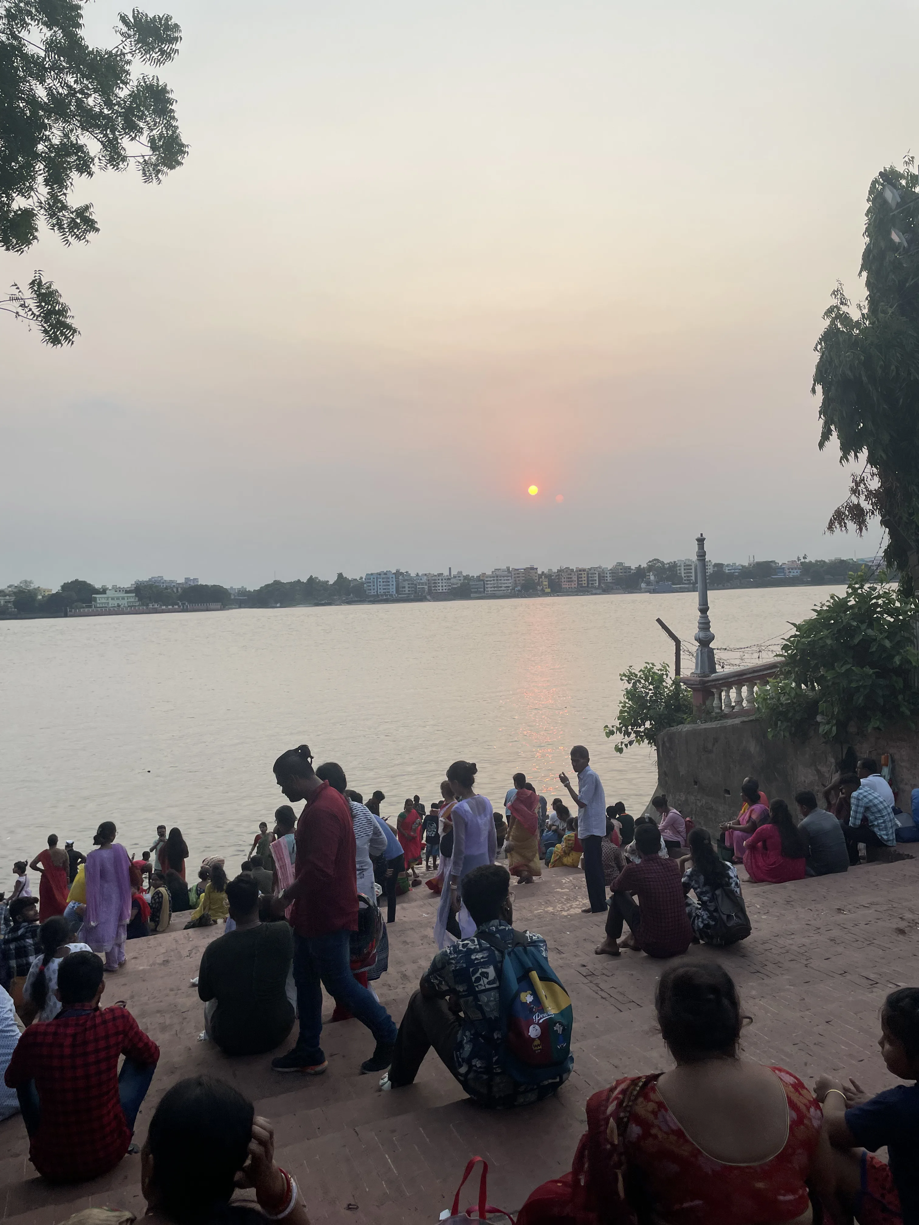 Multidão de pessoas reunida nos degraus de tijolos à margem do rio Hooghly, em Calcutá, contemplando o pôr do sol com o sol alaranjado refletindo nas águas.