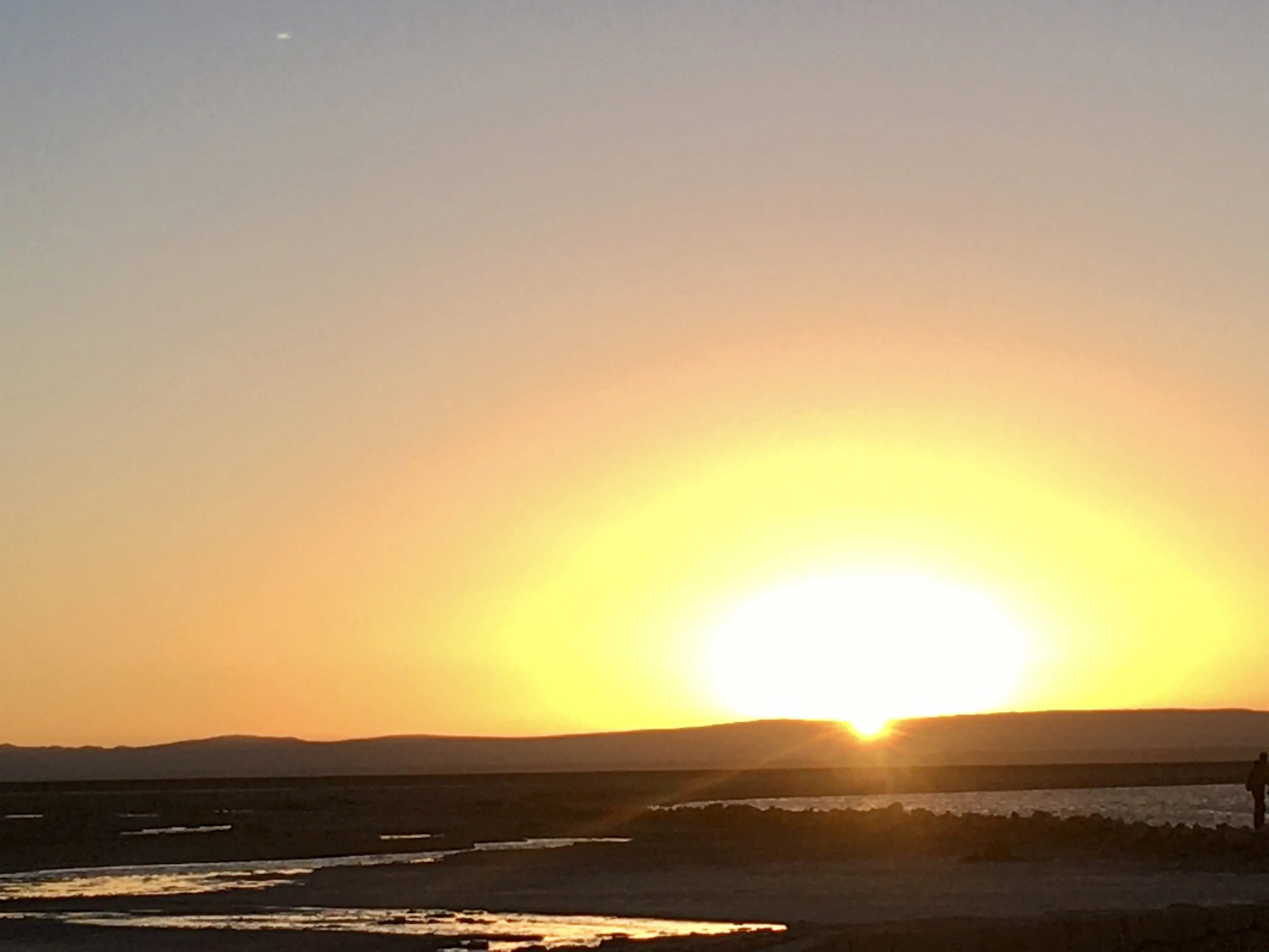 Sol brilhante e dourado se põe atrás das montanhas escuras iluminando a água no fim de tarde no Deserto do Atacama no Chile.