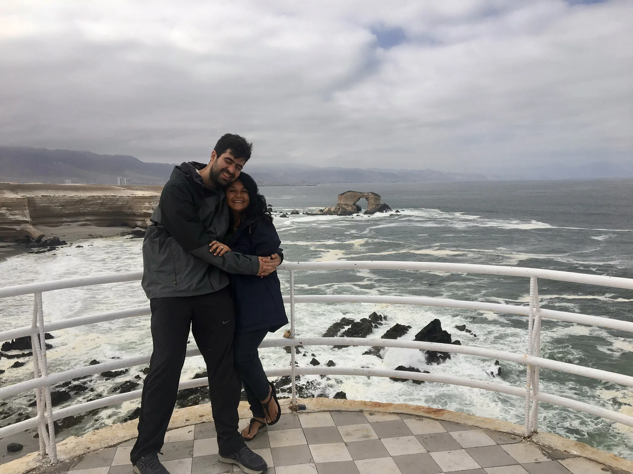 Casal de viajantes abraçados na grade do mirante do Monumento Natural La Portada, em Antofagasta, de costas para o famoso arco de pedra que forma um paredão íngreme esculpido pelo mar com um buraco no meio.