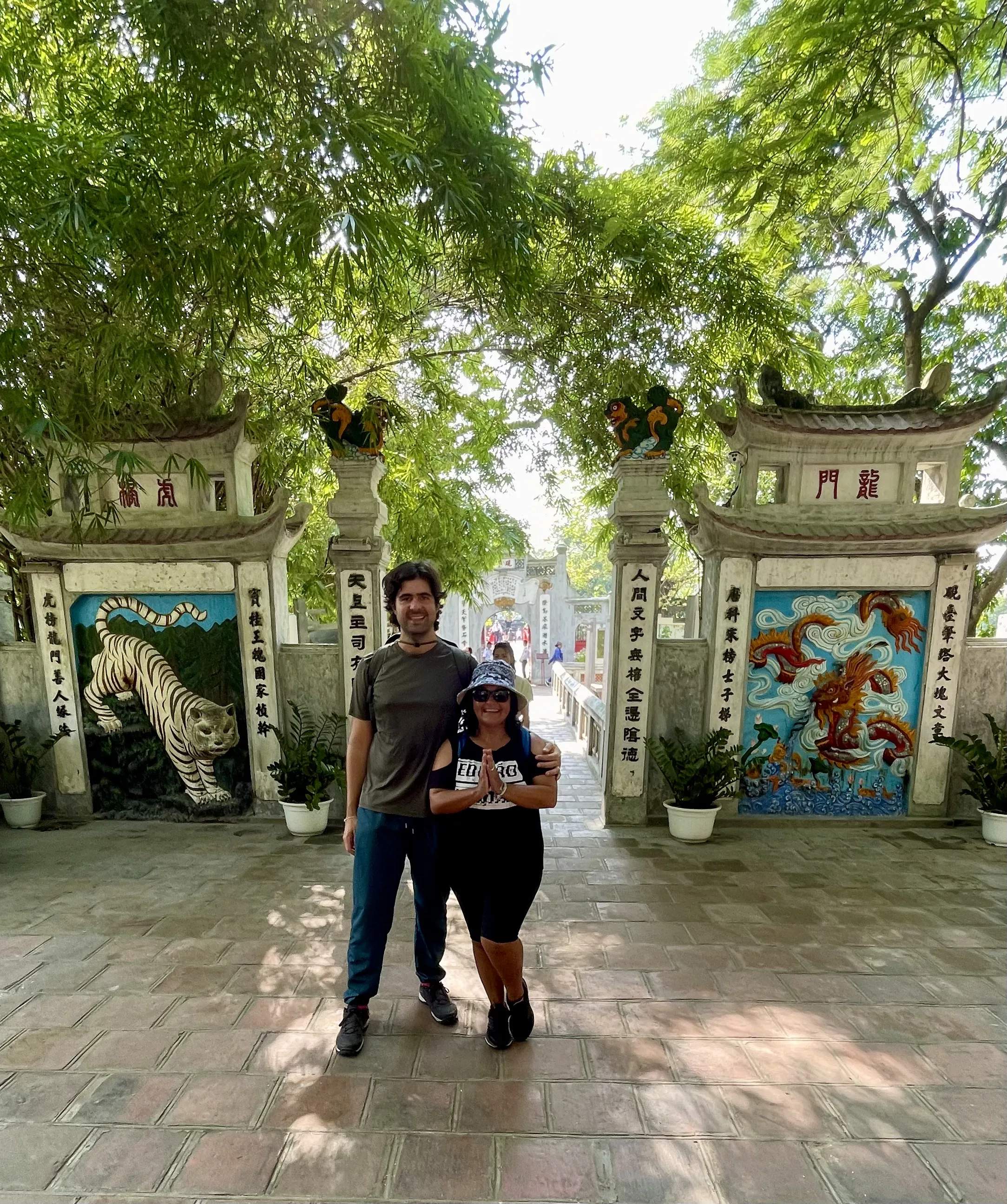 Casal posando de mãos juntas em gesto de respeito em frente ao portal de entrada do Templo Ngoc Son. O pórtico de alvenaria branca possui telhados cinzentos recurvados e painéis laterais emoldurados com relevos coloridos: à esquerda, um tigre branco com listras pretas pintado sobre fundo azul; à direita, um dragão vermelho sinuoso. Detalhes de árvores frondosas e bambus formam o dossel verde acima da estrutura.