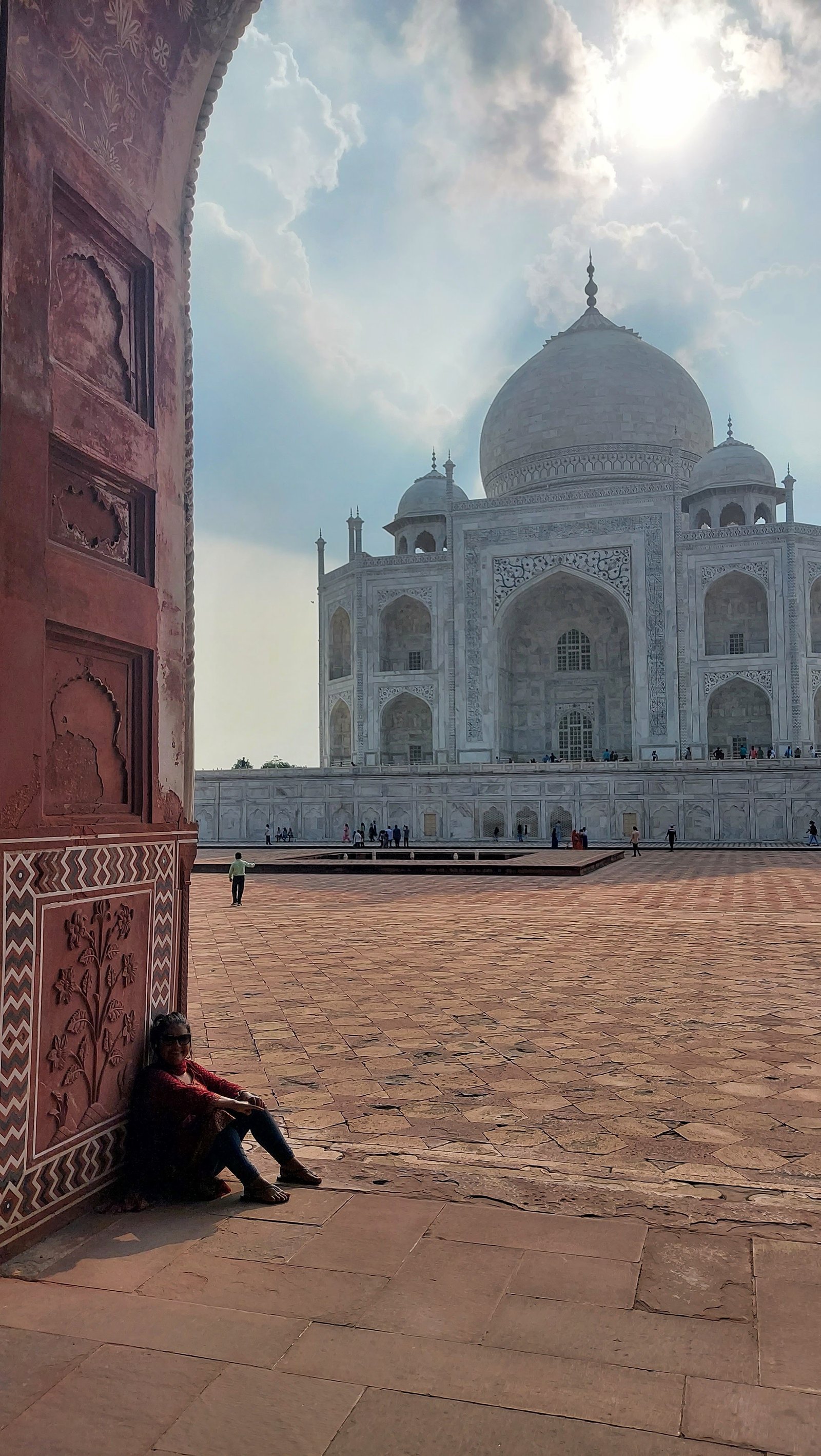 Visitante sentada no chão de arenito encostada em um pilar decorado com entalhes tradicionais de arenito vermelho, tendo ao fundo a grandiosa cúpula de mármore branco do Taj Mahal sob o sol.