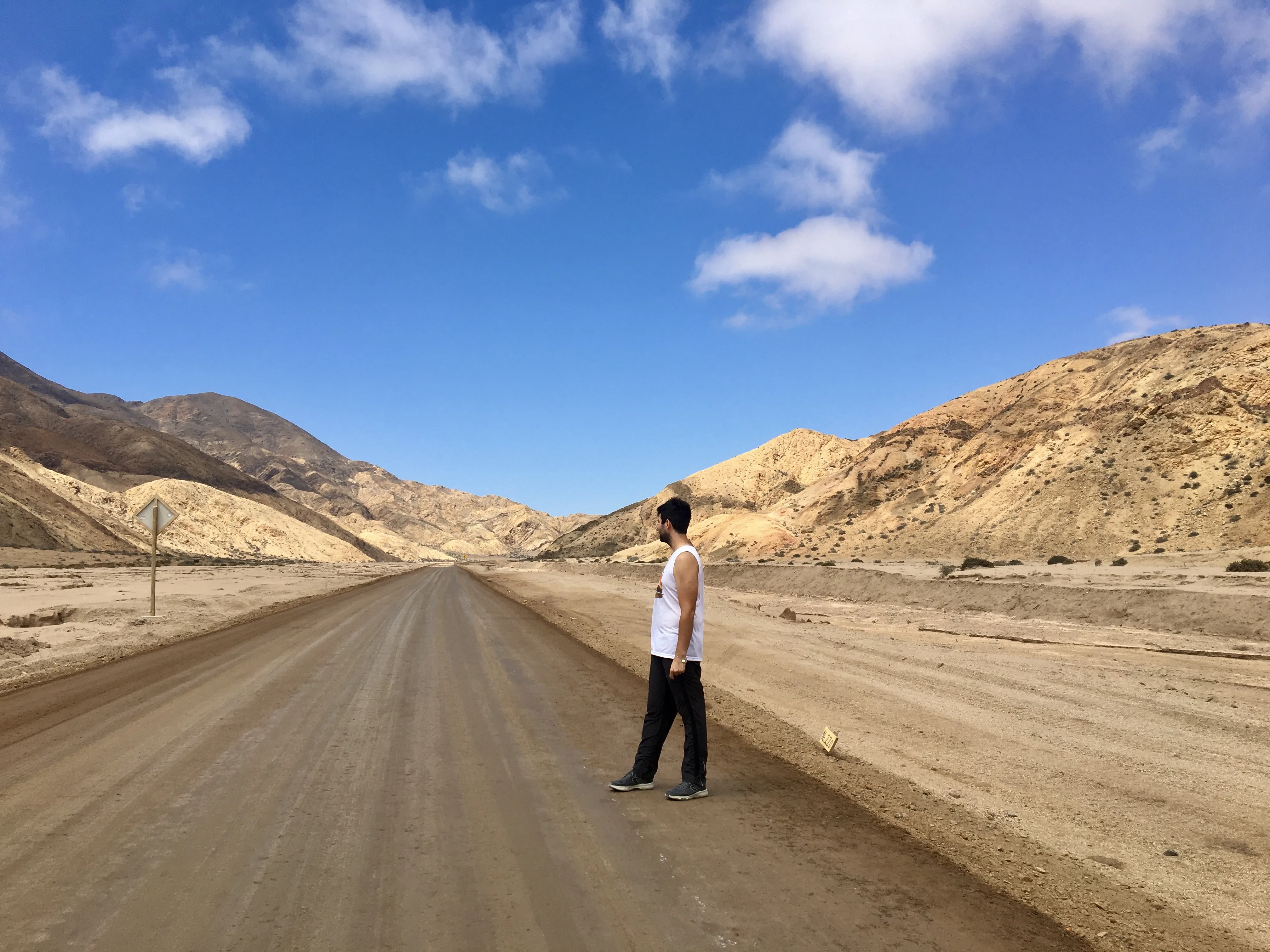 Homem em pé na ponta de uma estrada de asfalto vazia cortando a imensidão árida do Deserto do Atacama cercada por montanhas de terra batida na Quebrada de Chañaral.