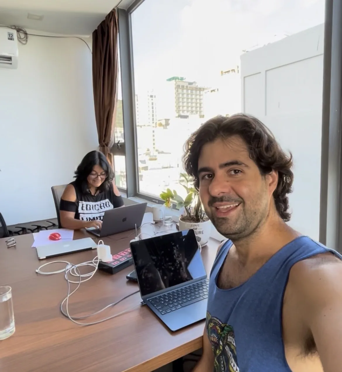 Pessoa posando sorridente em primeiro plano junto a uma mesa de reunião em um coworking em Da Nang, com uma segunda pessoa ao fundo trabalhando concentrada em um notebook cinza diante de uma grande janela envidraçada