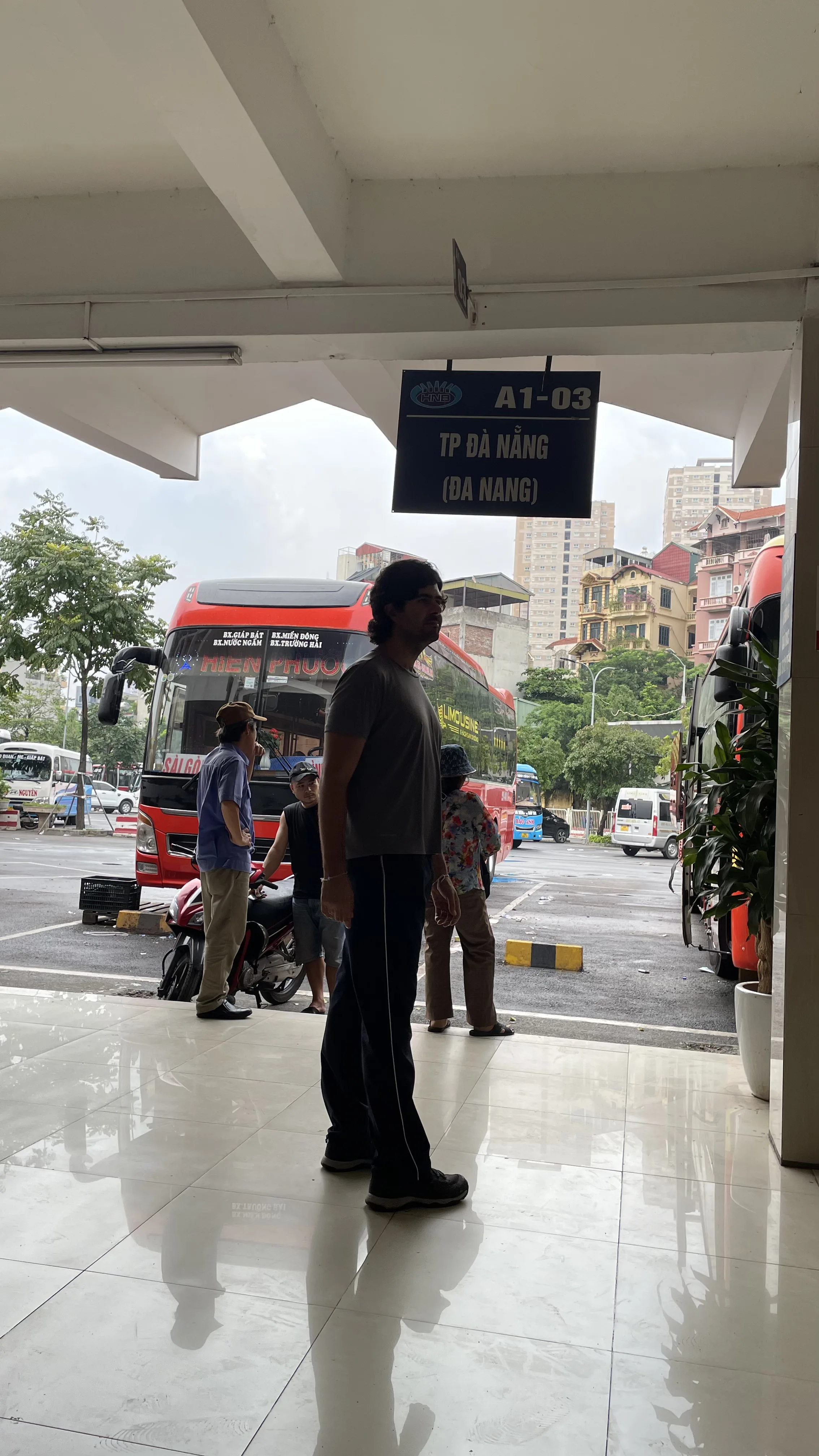 Homem em pé numa estação rodoviária movimentada em Hanói, com autocarros de viagem estacionados e placa indicativa de destinos ao fundo