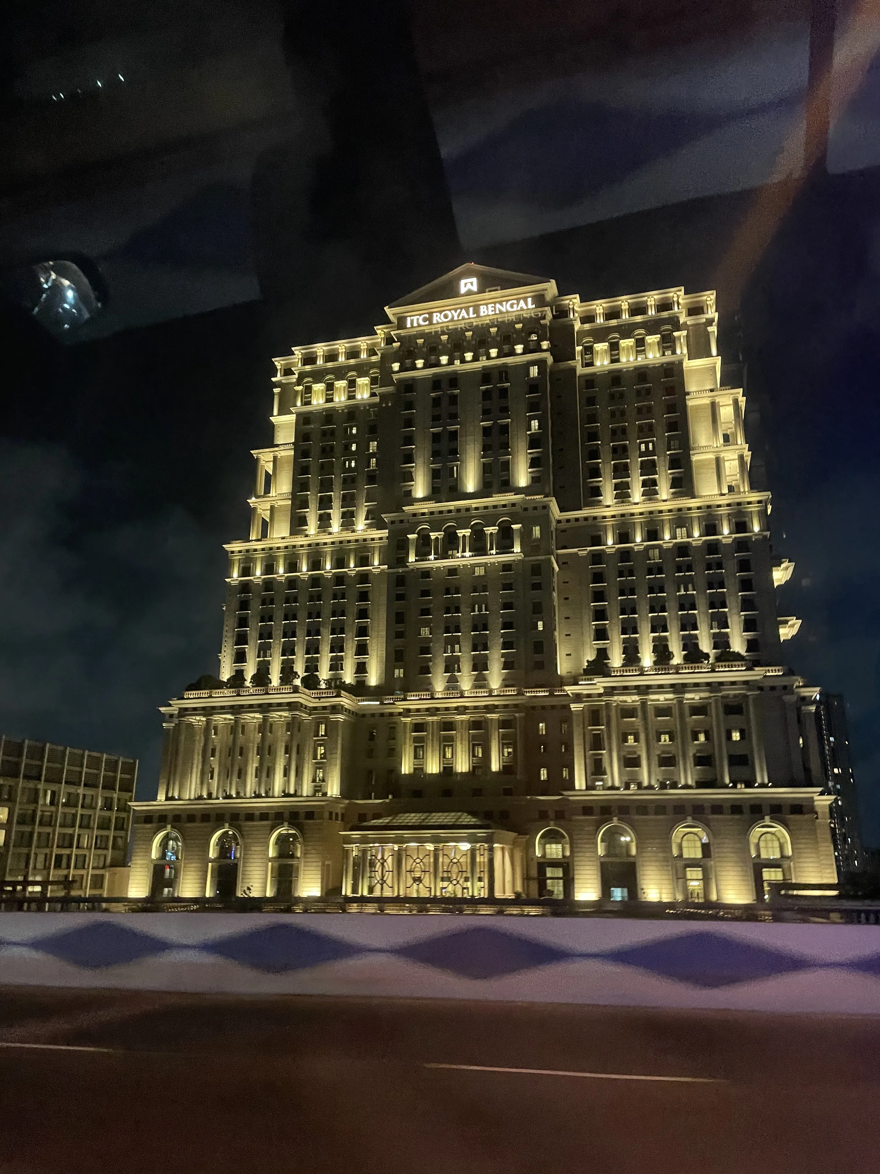 Fachada imponente do hotel ITC Royal Bengal em Calcutá, iluminada à noite com luzes amarelas em sua arquitetura clássica de múltiplos andares.