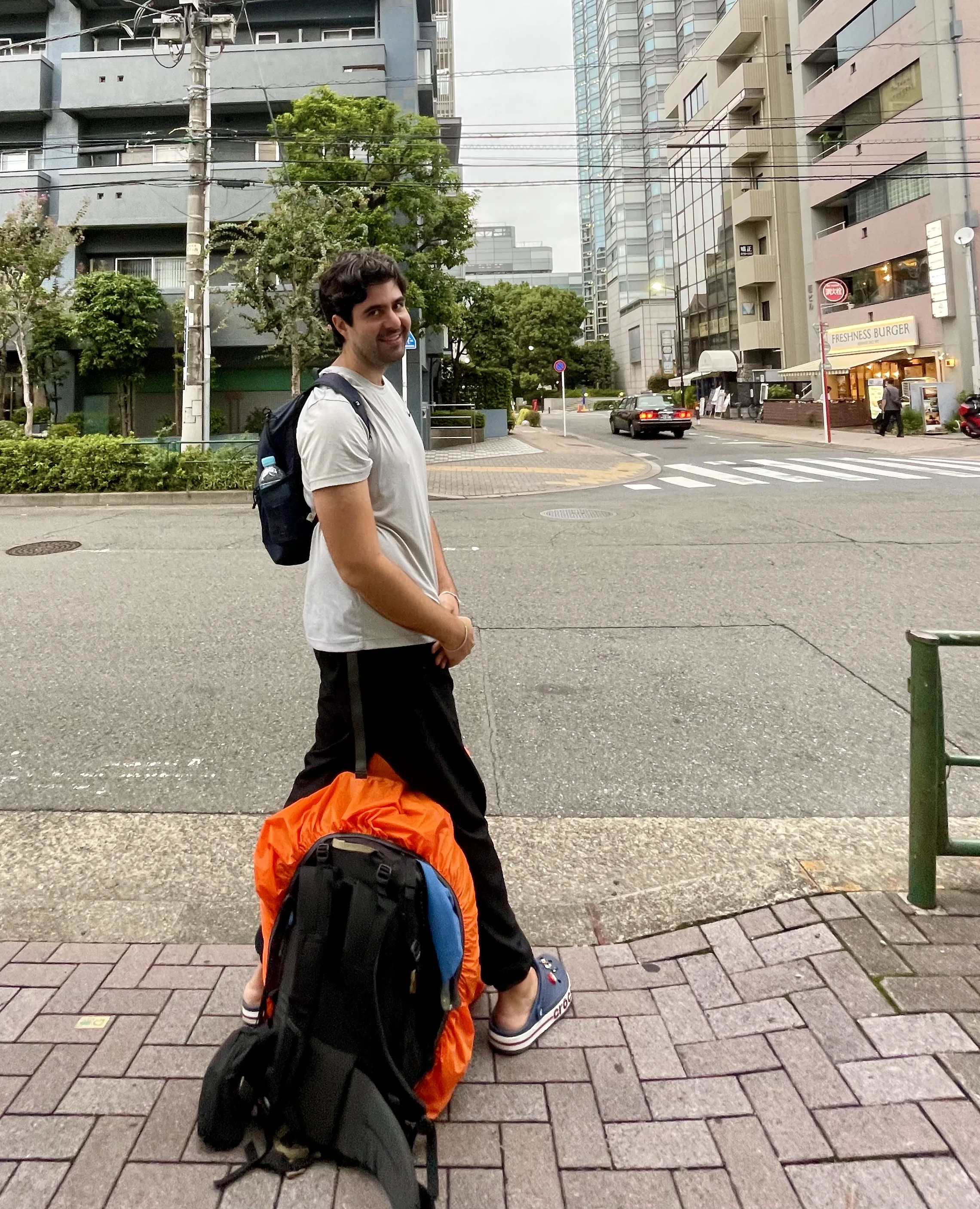 Pessoa em pé na calçada pavimentada de uma rua em Tóquio, com uma mochila preta nas costas e uma mala de viagem apoiada no chão equipada com capa de proteção laranja viva, tendo ao fundo edifícios urbanos, faixa de pedestres e um carro preto em trânsito.