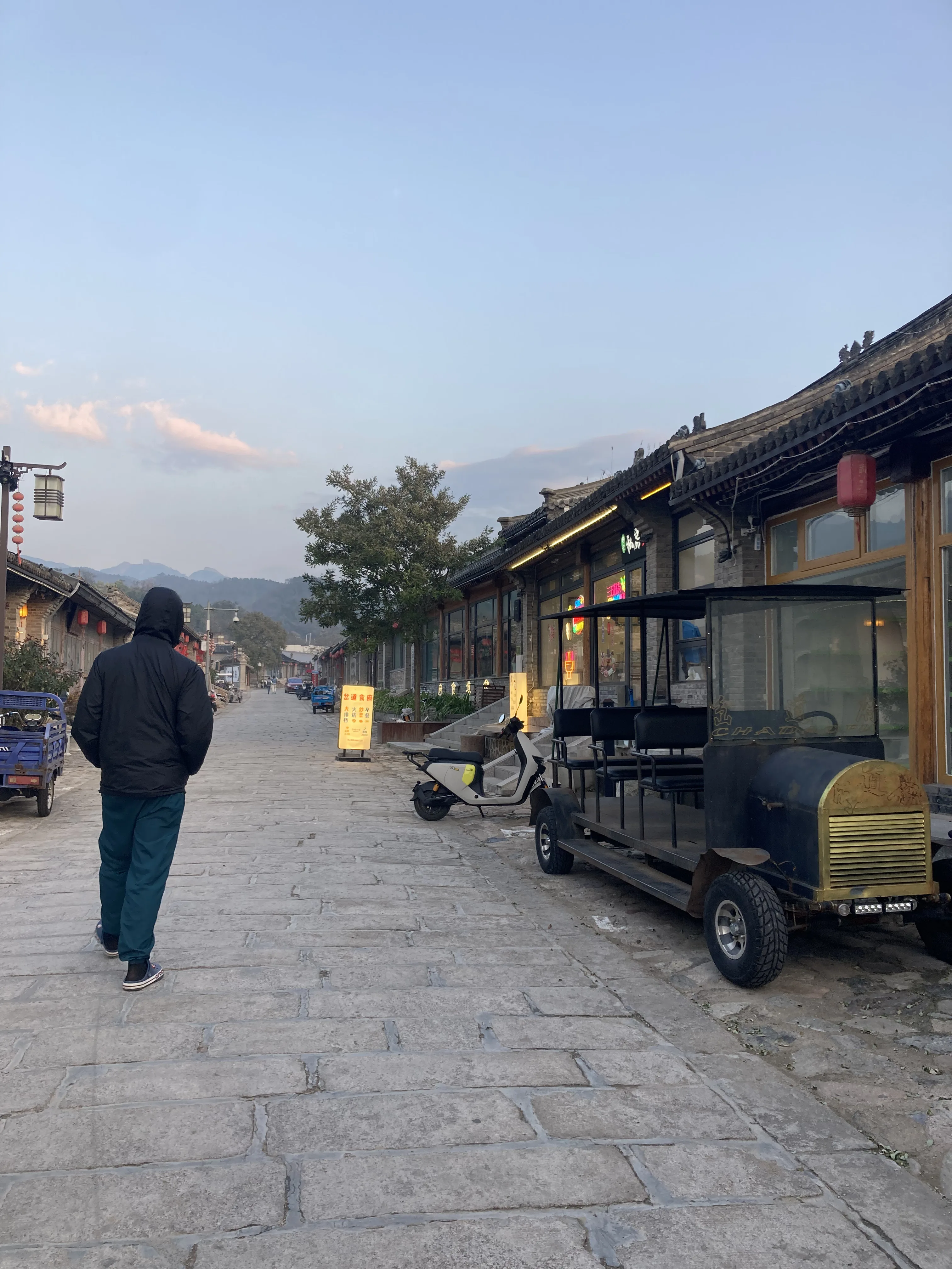 Homem de costas a caminhar por uma rua ampla de pedras pavimentadas na vila histórica de Badaling na China, ladeada por construções tradicionais, um carrinho elétrico antigo de turismo estacionado à direita e montanhas ao fundo.