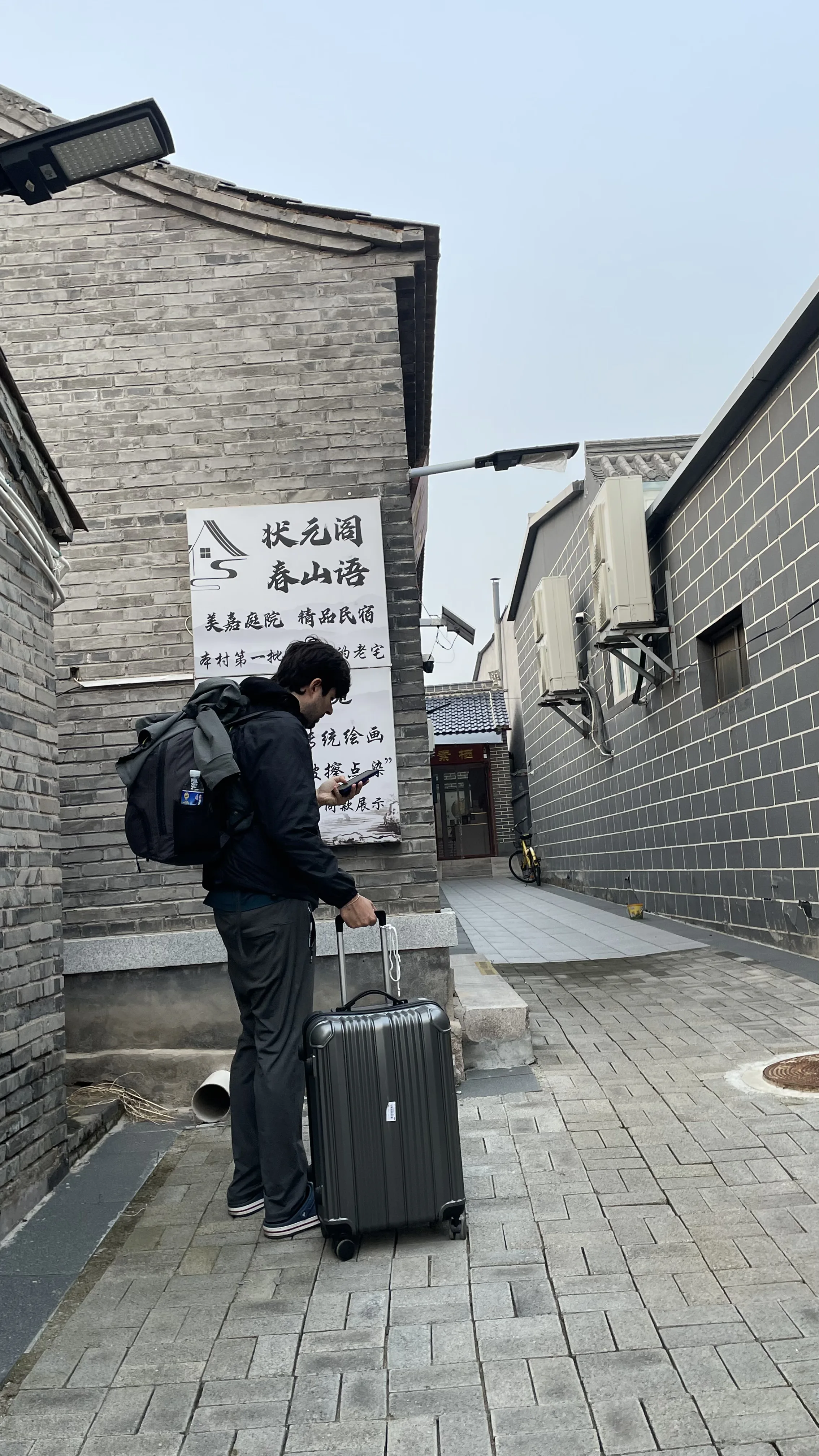 Turista com mochila e mala de rodinhas conferindo o celular em uma rua estreita de piso de pedra entre casas tradicionais de tijolos cinzas em vila histórica perto de Badaling.