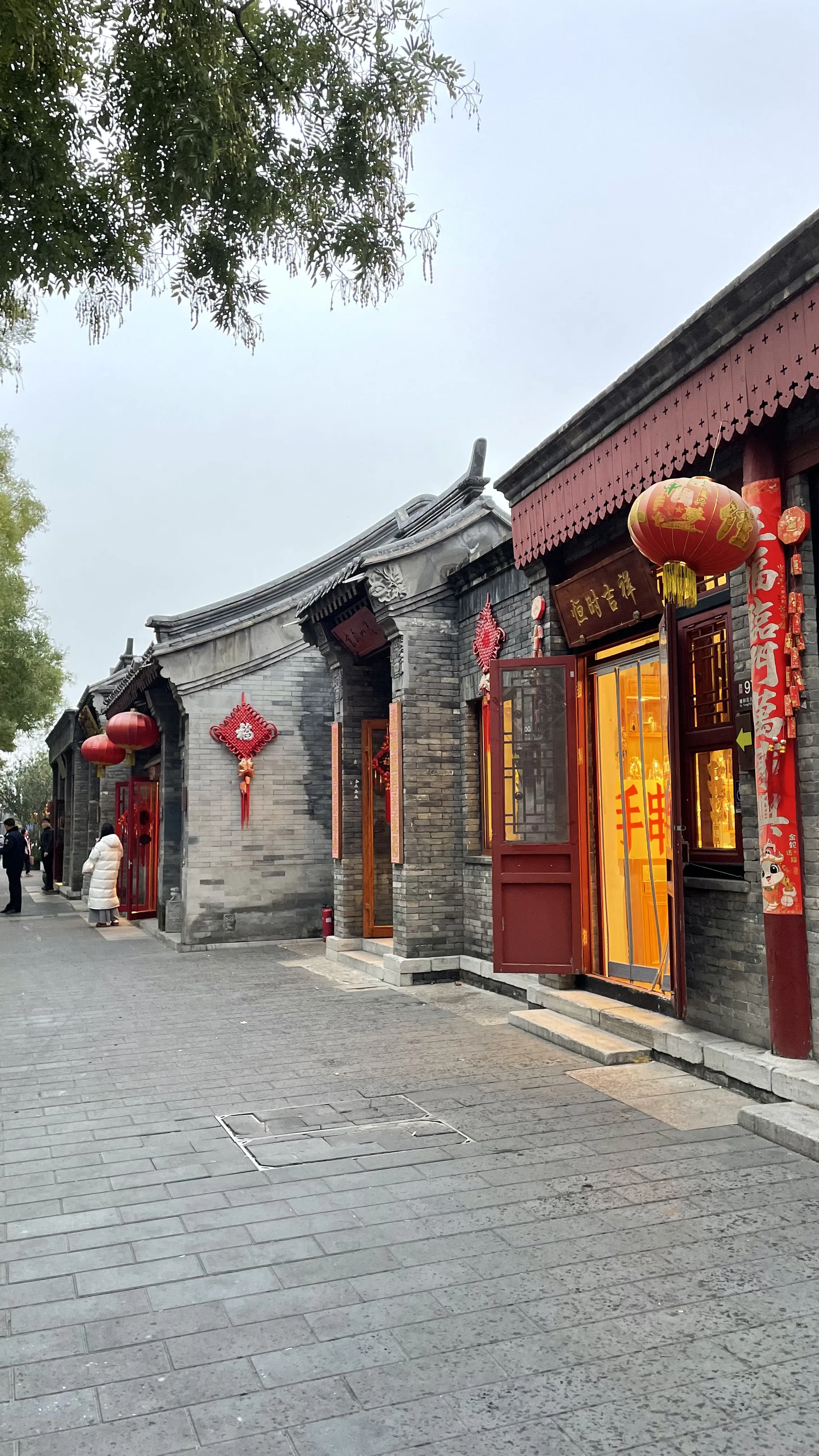 Rua tradicional em beco hutong em Pequim com construções de tijolos cinzentos, portas vermelhas, lanternas e decorações festivas.