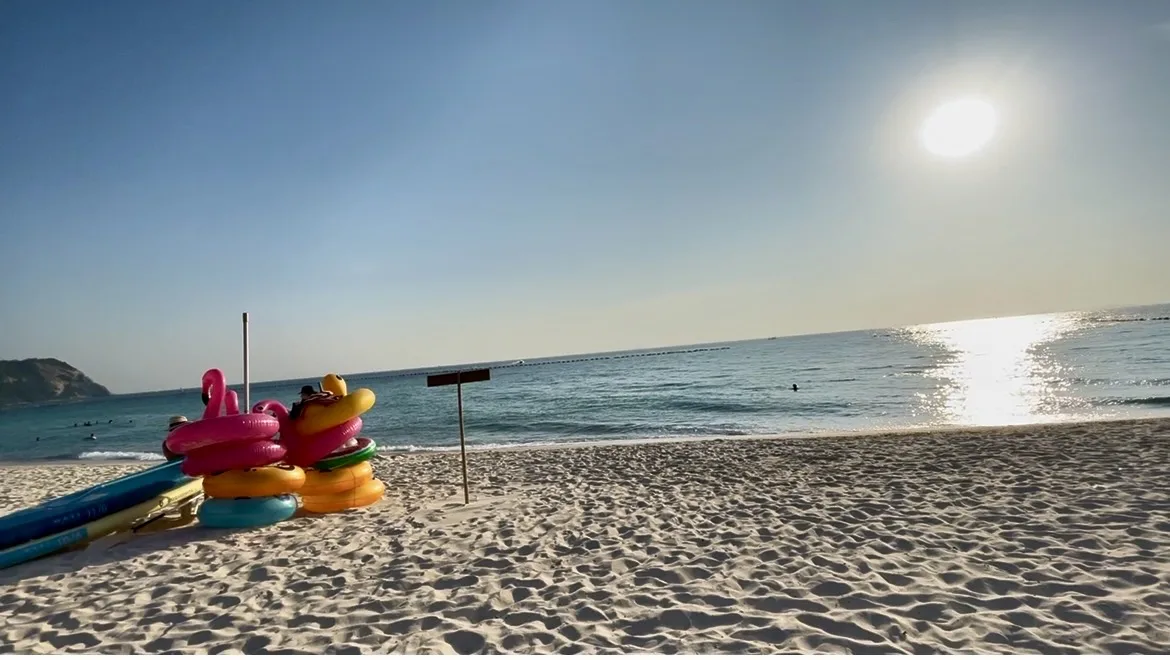 Bóias infláveis coloridas empilhadas na areia branca da praia em Ko Lan, com o mar e o sol brilhando ao fundo.