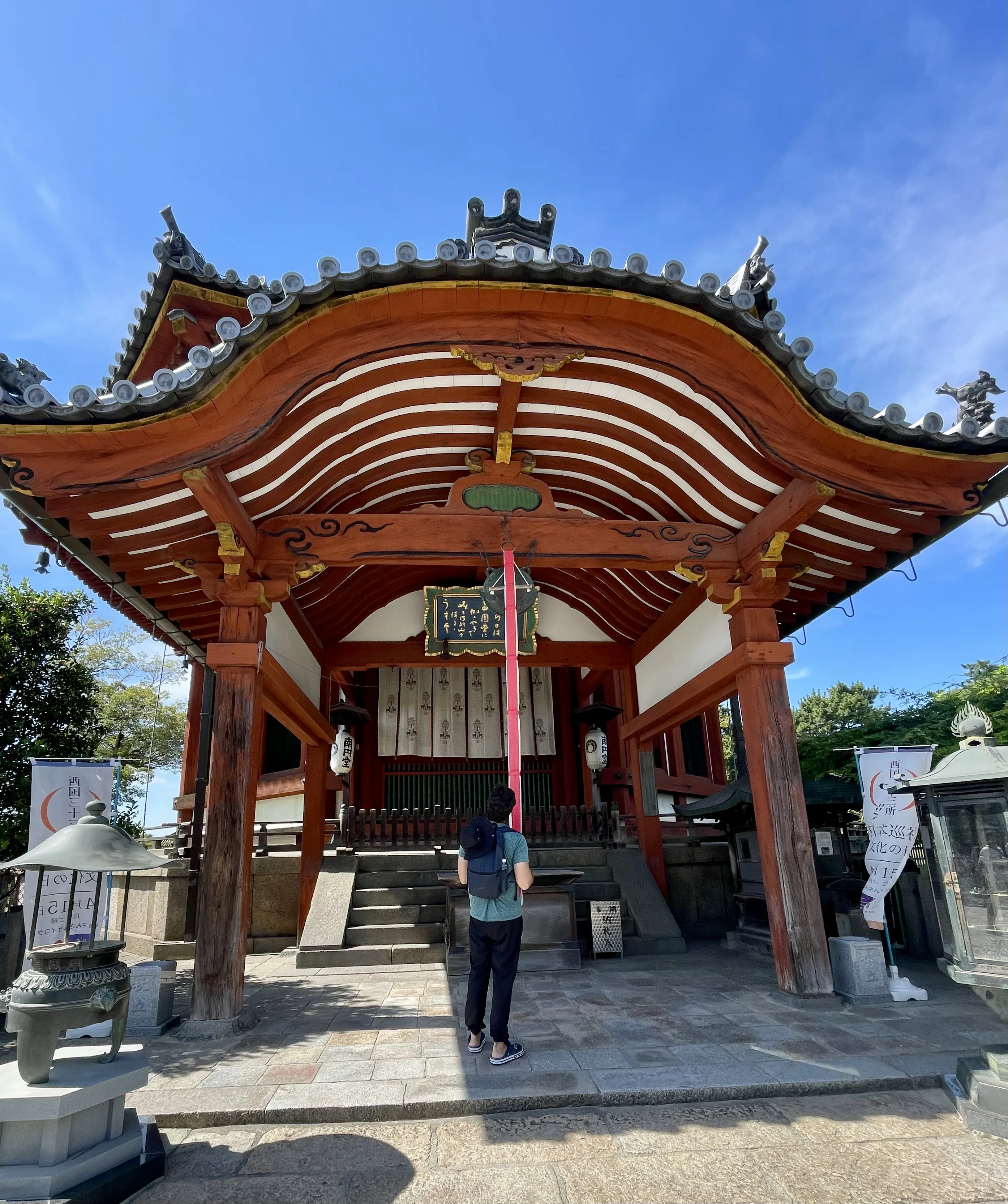 Homem de costas com mochila puxa a corda vermelha para tocar a sineta em altar tradicional de templo sob céu azul em Nara no Japão.